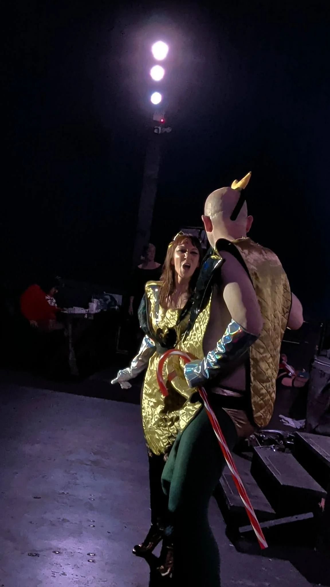 Nightclub scene with a woman dressed in shiny gold and silver costume dancing or performing. She is facing a man in a costume with a large unicorn horn. The dark background has some equipment and another person in red. Stage lighting illuminates the scene.