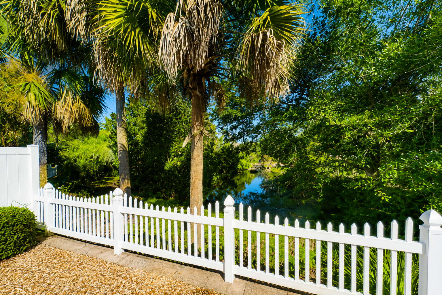 Peaceful backyard view at Sunshine Stay Creekside with white picket fence, mature palms, and the creek beyond.