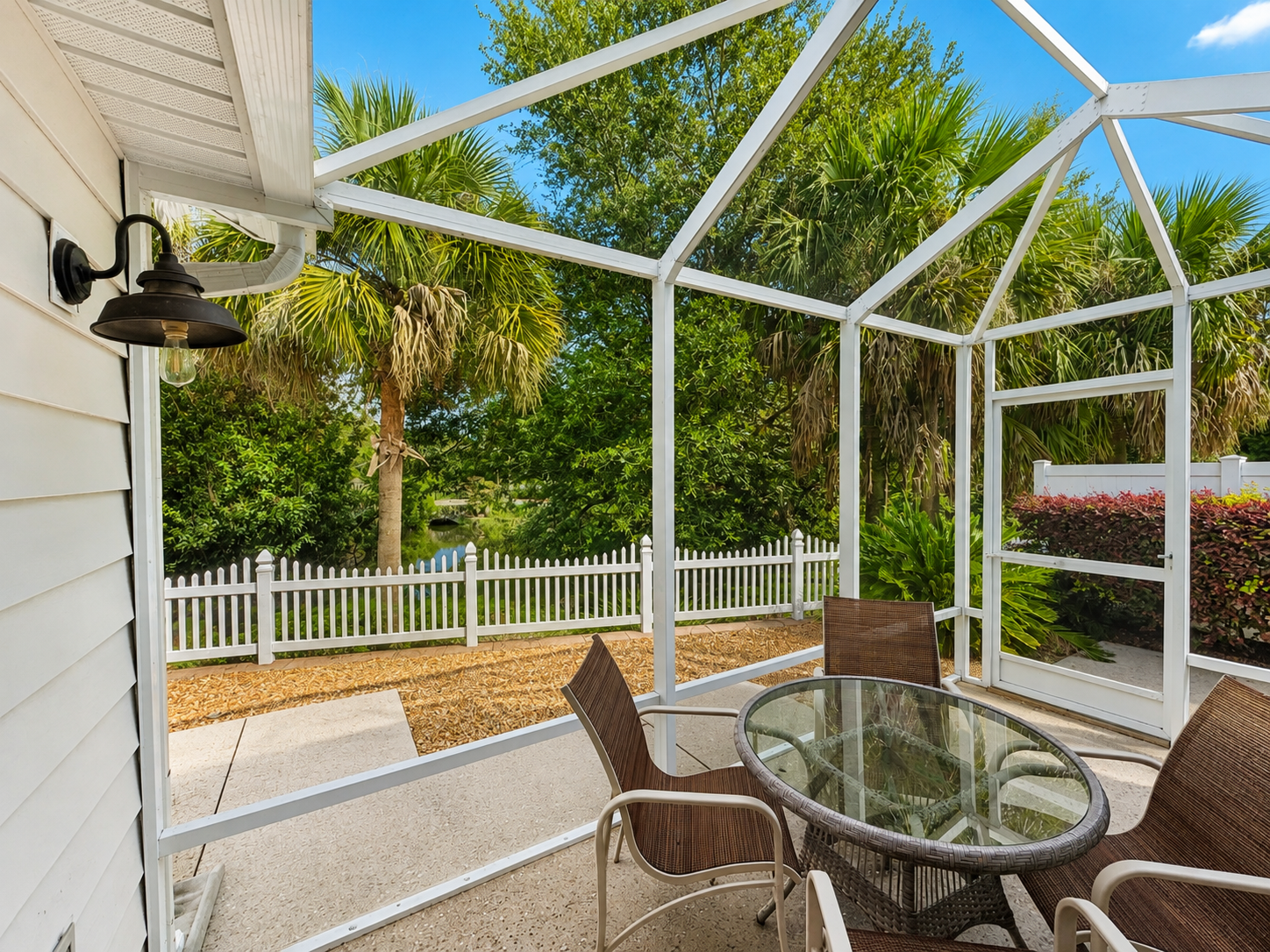 Screened lanai at Sunshine Stay Creekside with outdoor dining and views of mature palms, greenery, and the white picket fence beyond.