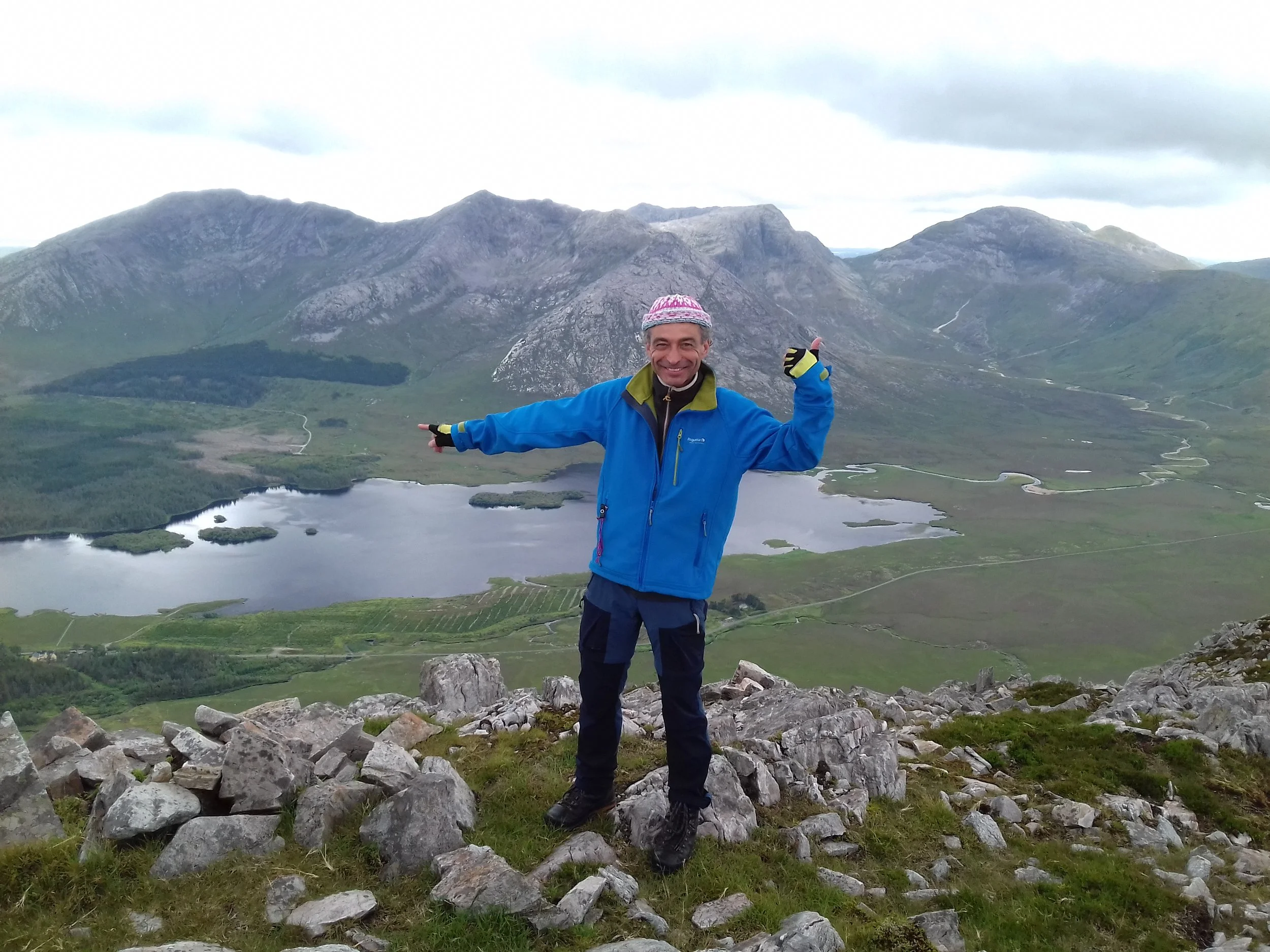 A man standing on a rocky hilltop in outdoor gear, smiling and pointing with both hands, set against a scenic mountain and lake landscape under cloudy skies.
