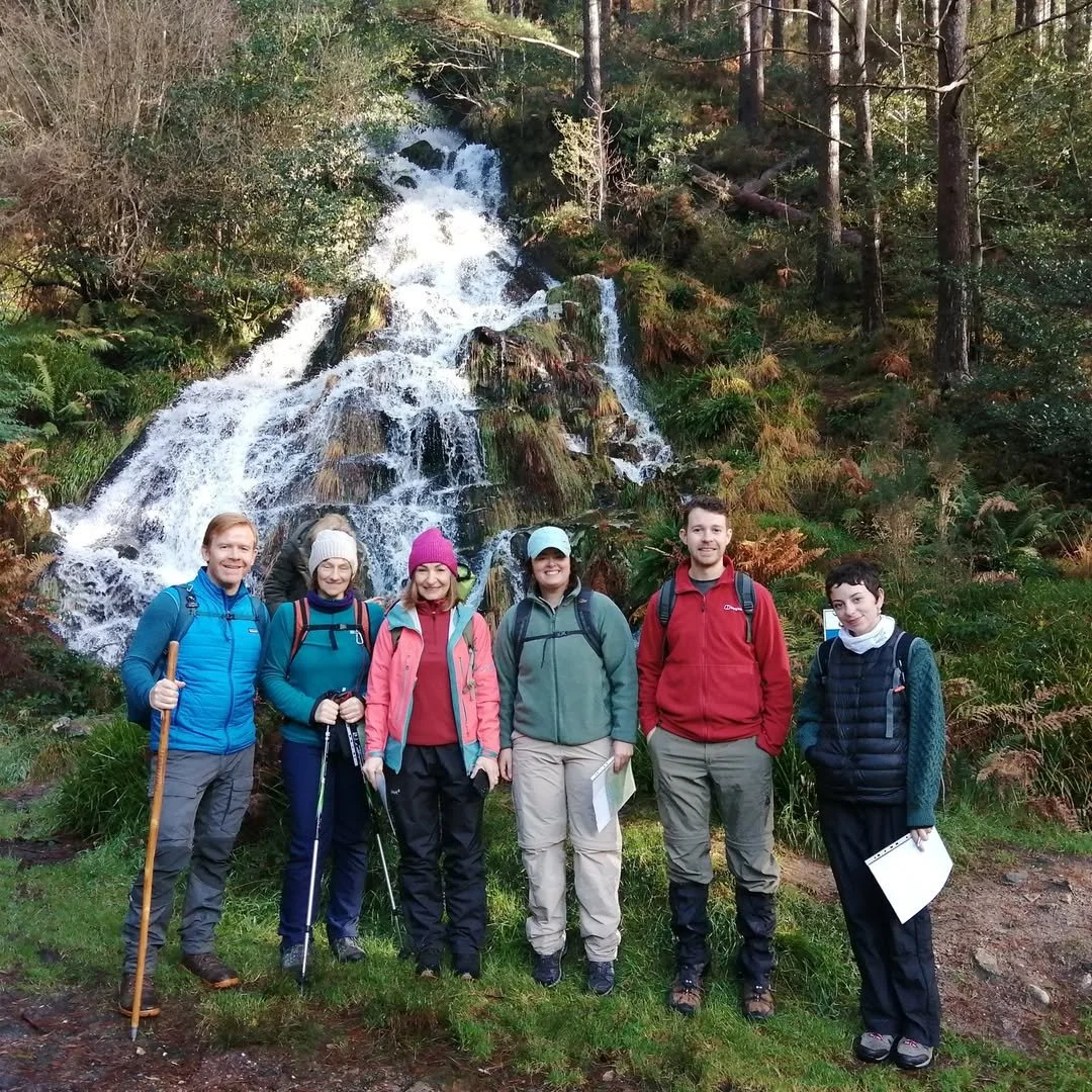 Group of six hikers standing in front of a waterfall in a forest, dressed in outdoor gear and holding trekking poles.