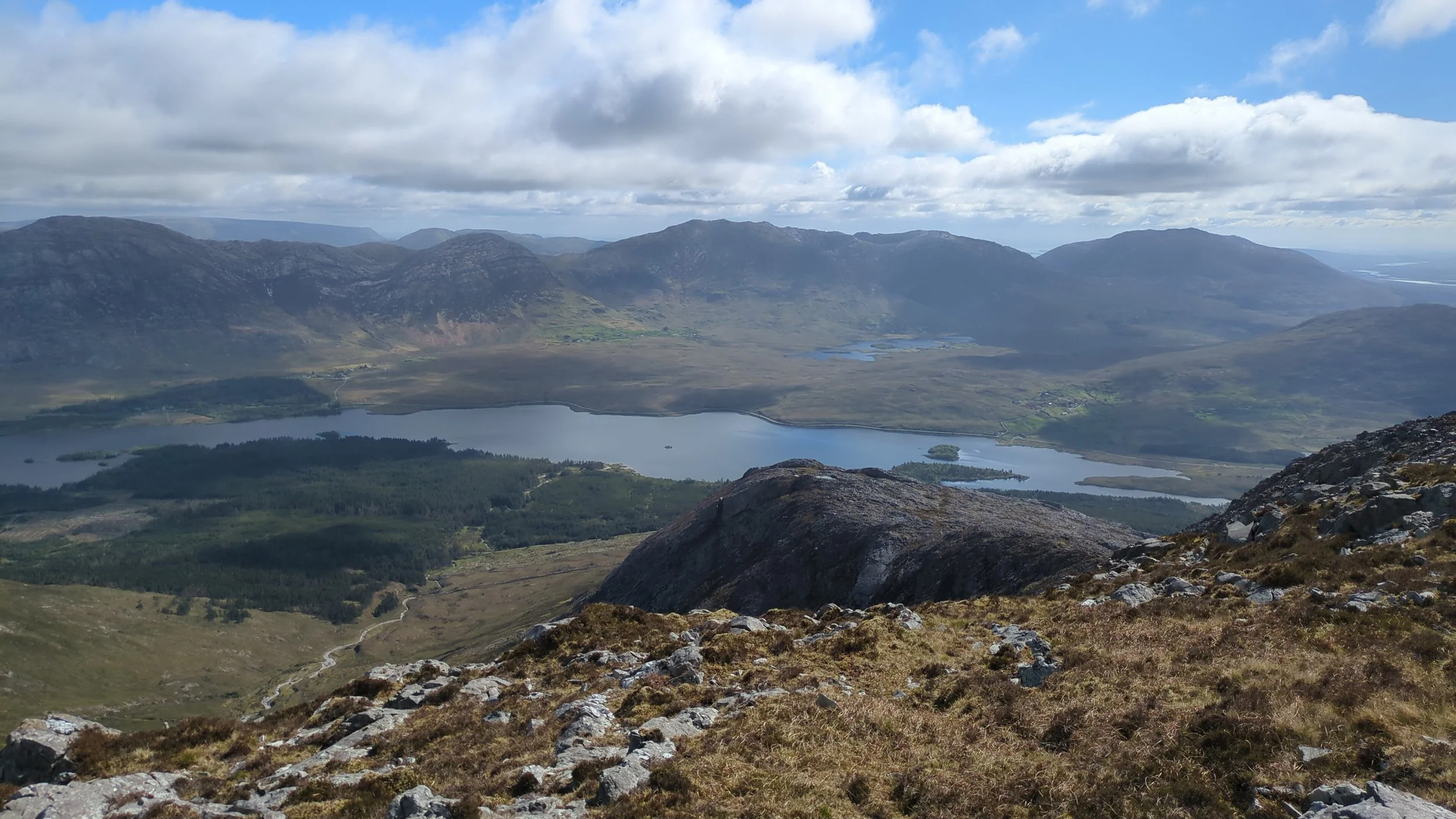 A scenic view of a mountain landscape with a lake in the valley, surrounded by green and rocky terrain under a partly cloudy sky.