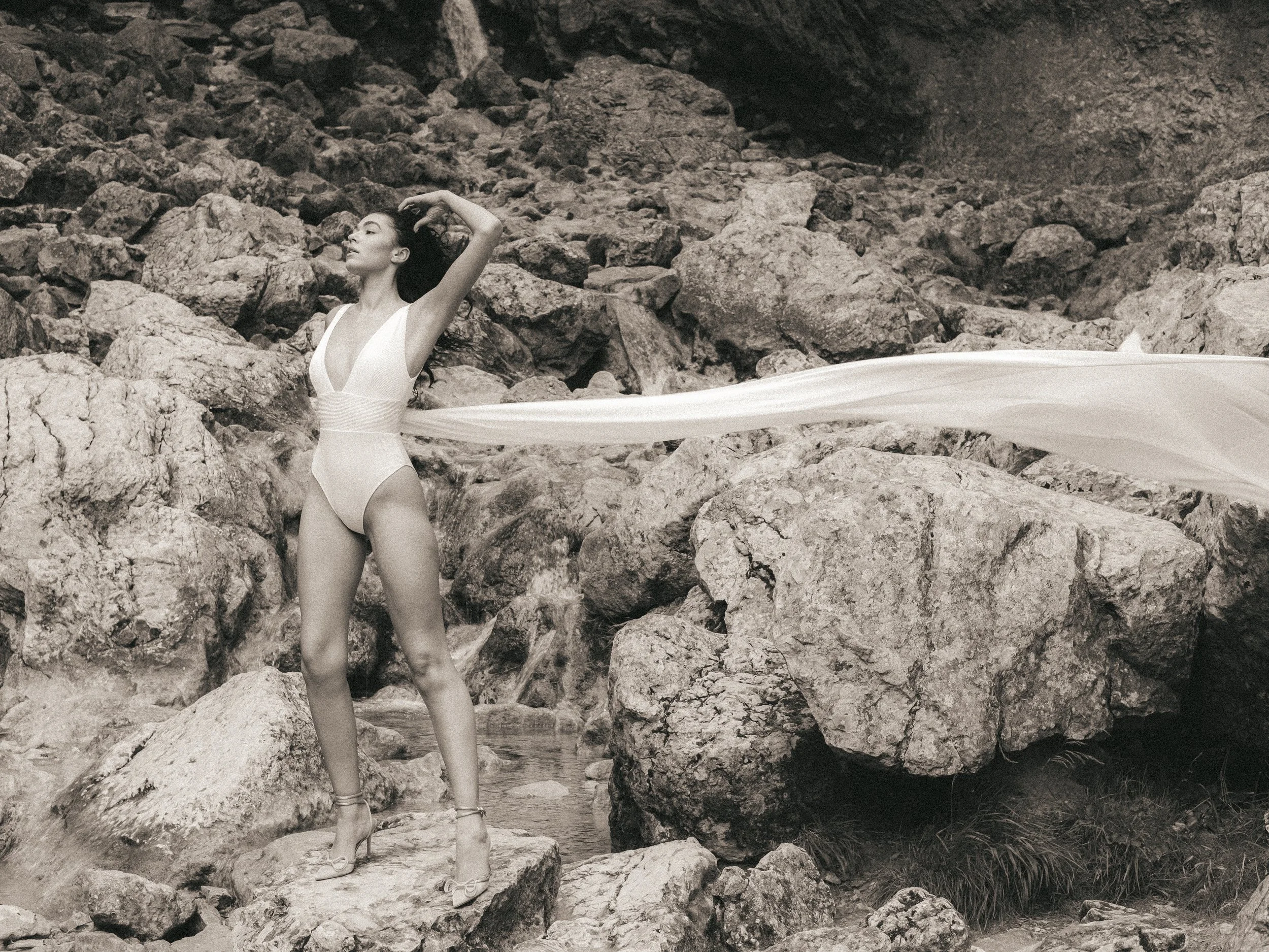 A woman in a white swimsuit standing on rocks near a small body of water, with her right arm raised and her hair flowing, against a background of large rocks and a cave.