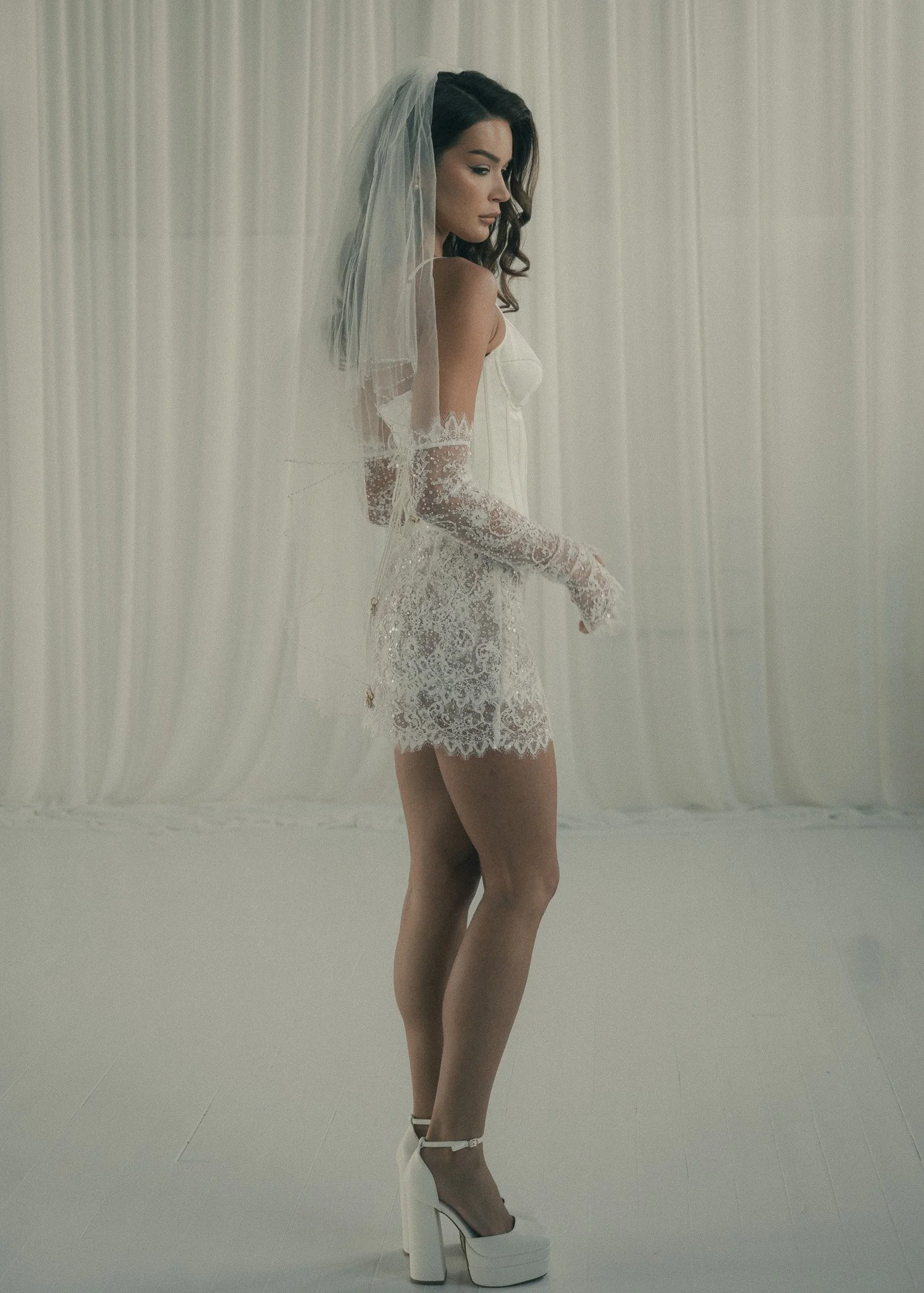A woman in white lace lingerie and white high-heeled platform shoes standing behind a sheer white curtain.