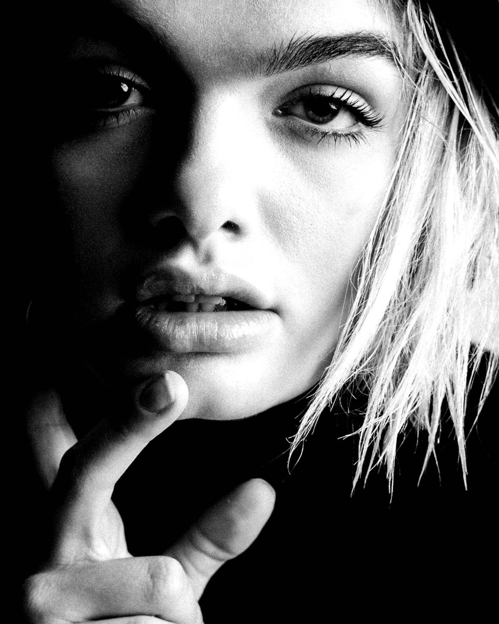 A close-up black and white photograph of a woman's face focusing on her eye and lips, with her hair partially visible and her hand near her face.