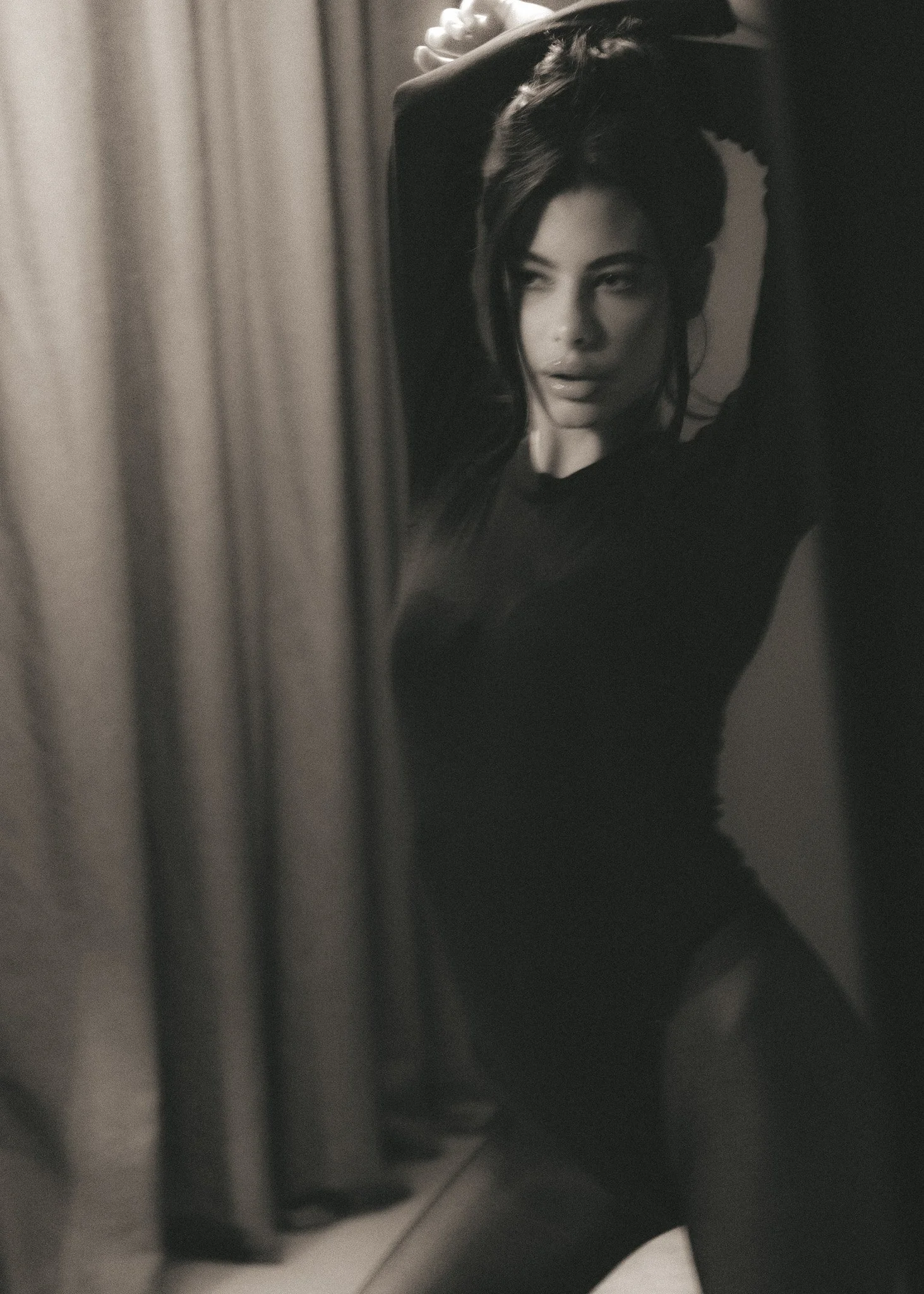 Black and white photo of a woman with long hair and a serious expression, looking into a mirror, with her arm raised and hand on her head.