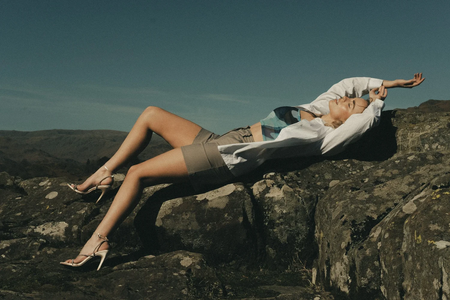A woman reclining on rocks outdoors, wearing a white shirt, tan shorts, and high heels, with her eyes closed and arms raised above her head against a clear sky.