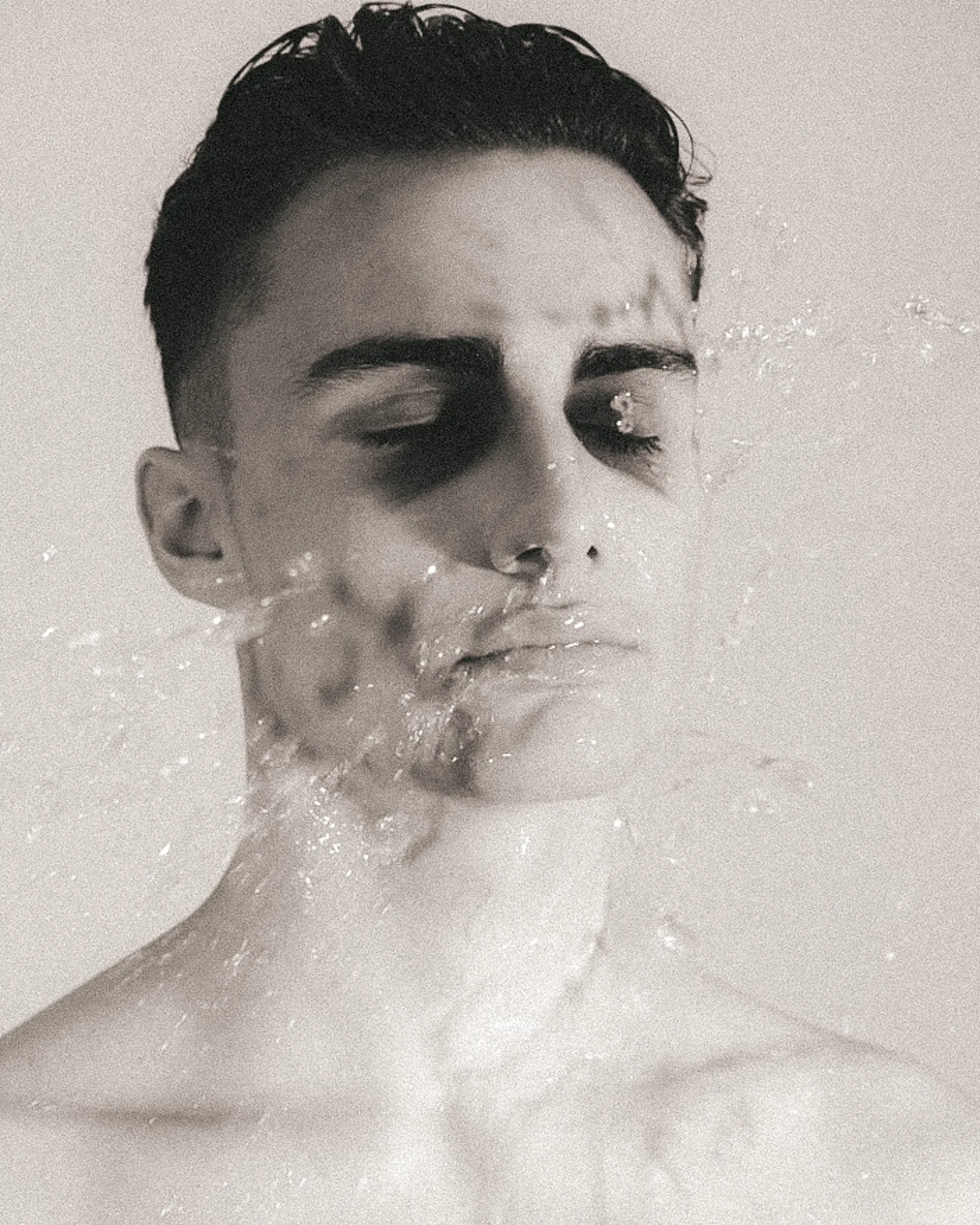 A black and white close-up photo of a young man's face with wet skin, slicked-back hair, closed eyes, and water droplets on his face and neck.
