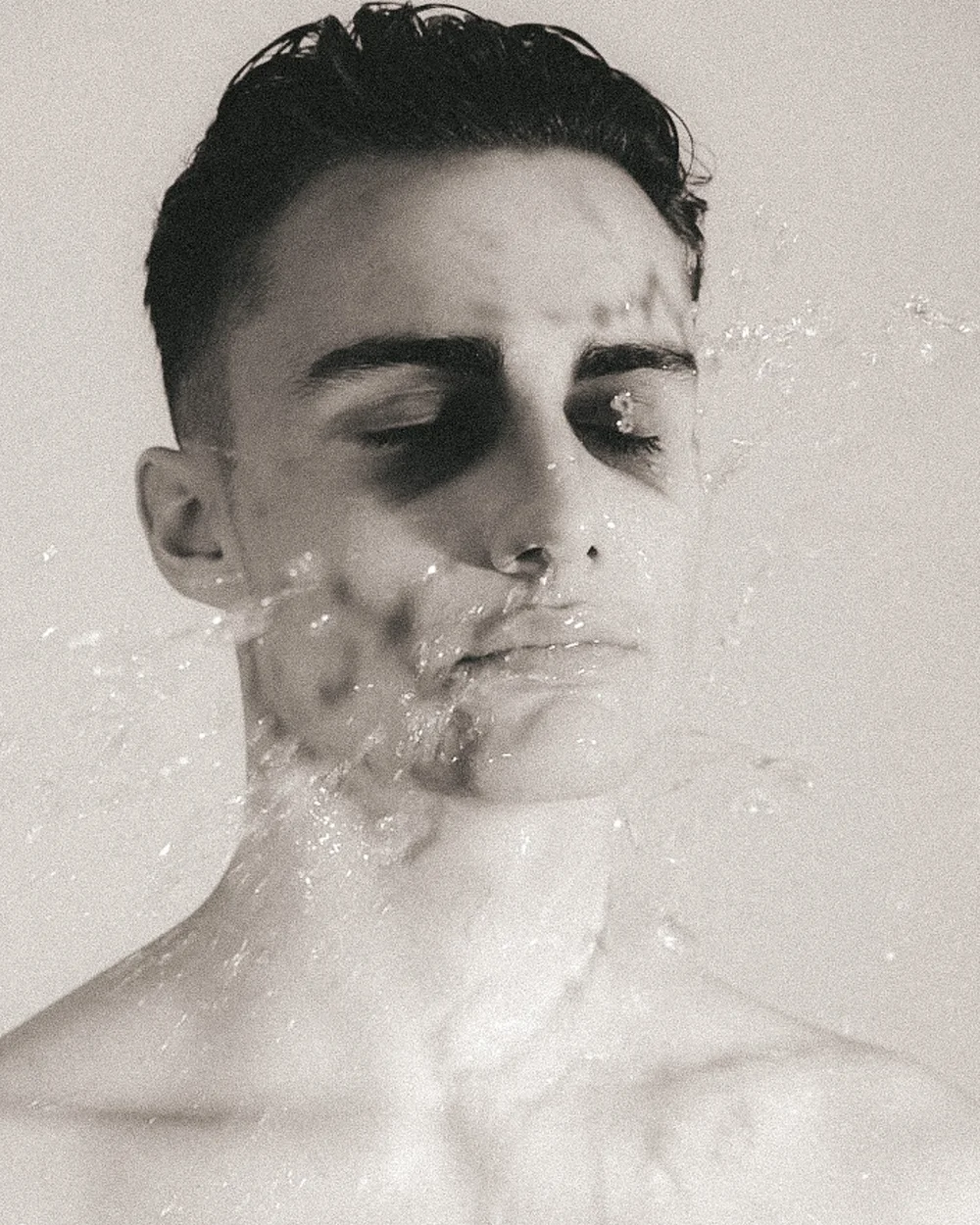 A black and white close-up photo of a young man's face with wet skin, slicked-back hair, closed eyes, and water droplets on his face and neck.