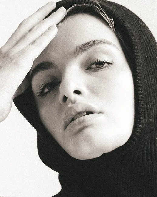 Close-up black and white portrait of a woman wearing a hooded garment, with her hand on her forehead, looking directly at the camera with a relaxed expression.