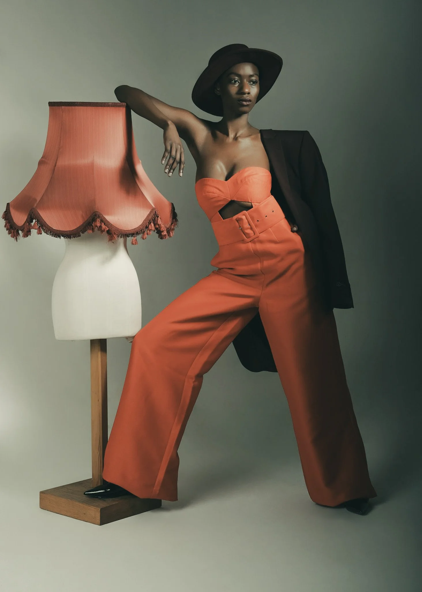 A woman dressed in an orange strapless top and matching wide-leg pants, wearing a black wide-brimmed hat, poses with her left arm resting on an orange lampshade on a white mannequin and slightly leaning to the right, with a black blazer draped over h