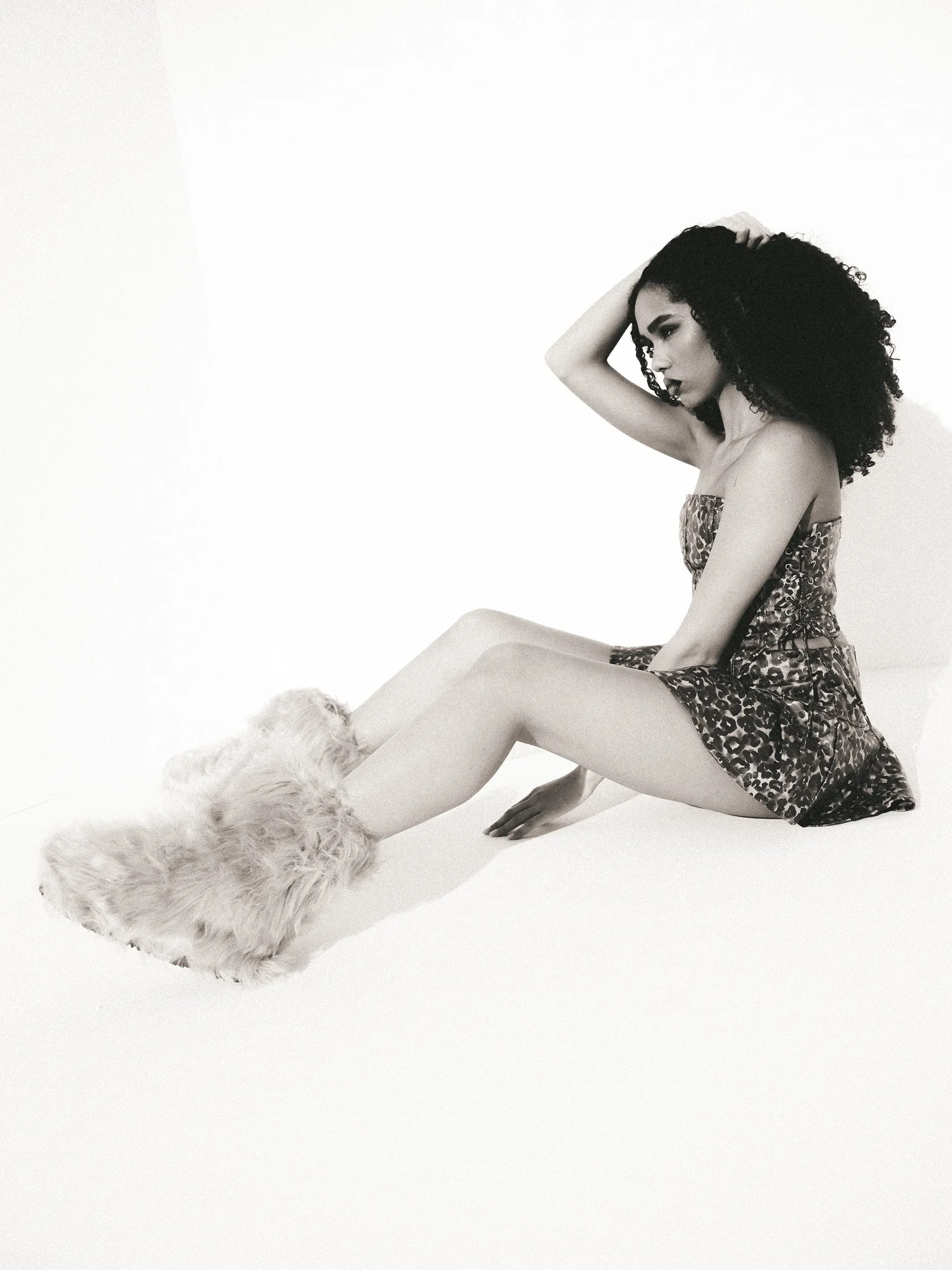 Black and white photo of a woman with curly hair sitting on the floor with her legs extended. She is wearing a leopard print dress and furry slippers, with one hand on her head and the other resting on the floor.