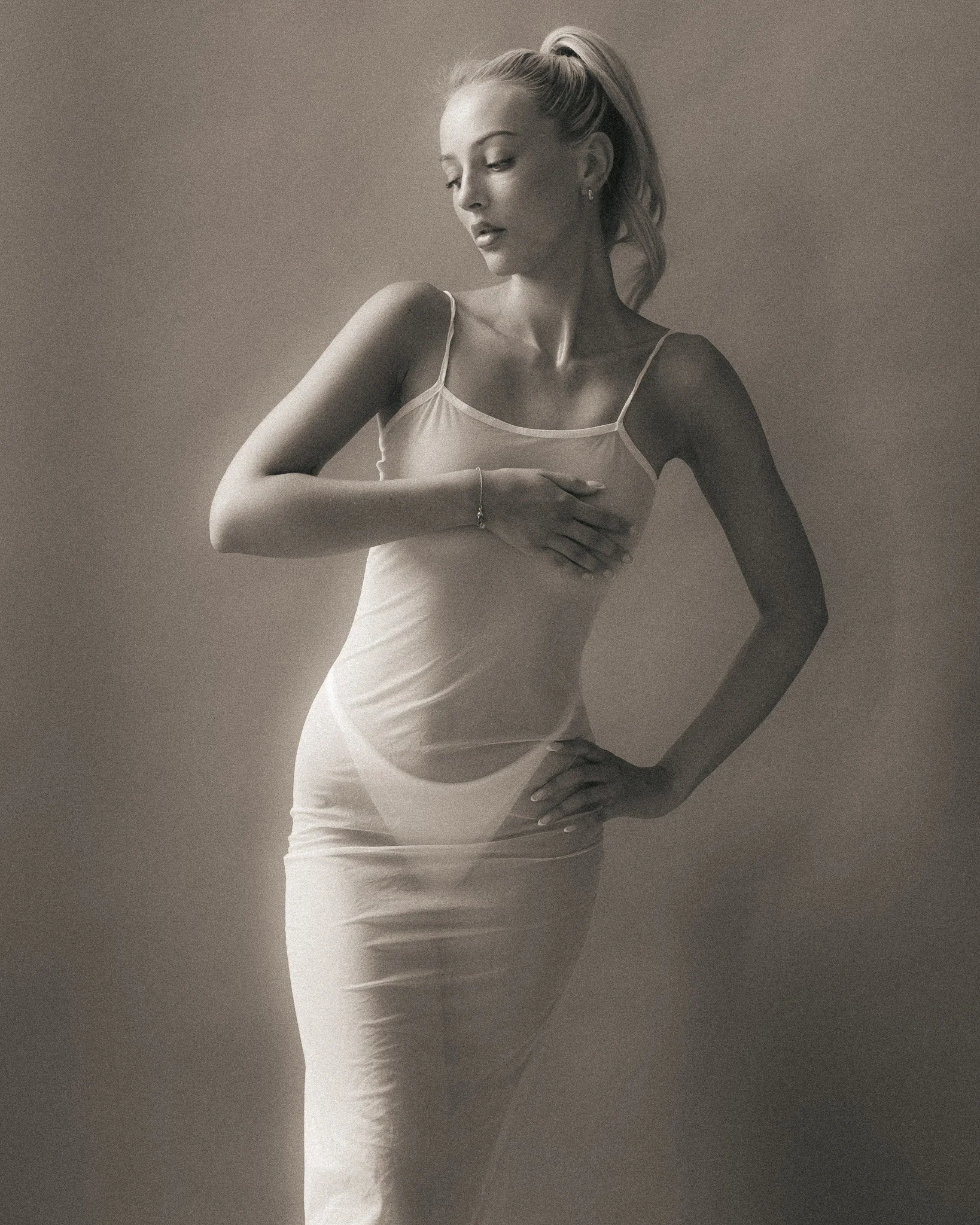 Black and white photograph of a woman in a fitted dress, standing sideways with her head slightly tilted down and her left arm crossing her chest. Her hair is pulled back in a high ponytail.