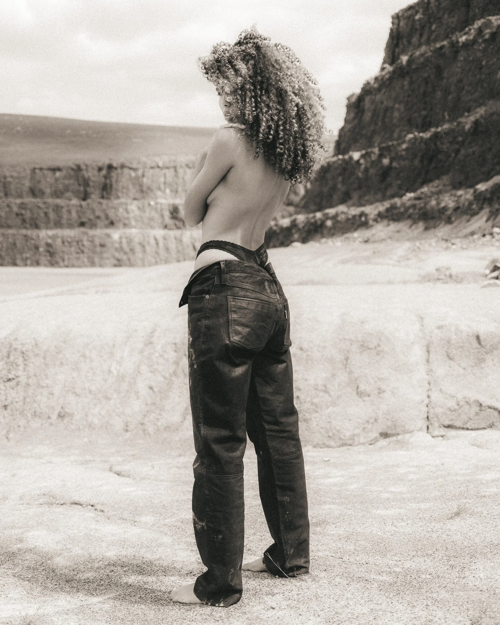 A woman with curly hair standing on a sandy beach, facing sideways with arms crossed, wearing black pants and no top, with a rocky cliff in the background.