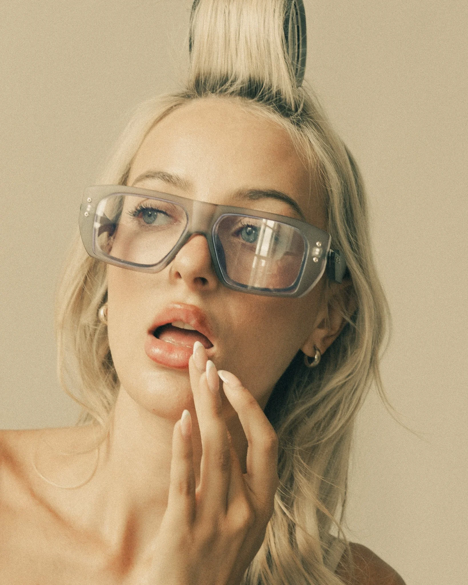 A woman with blonde hair wearing oversized sunglasses, with her tongue out and finger near her lips, against a beige background.