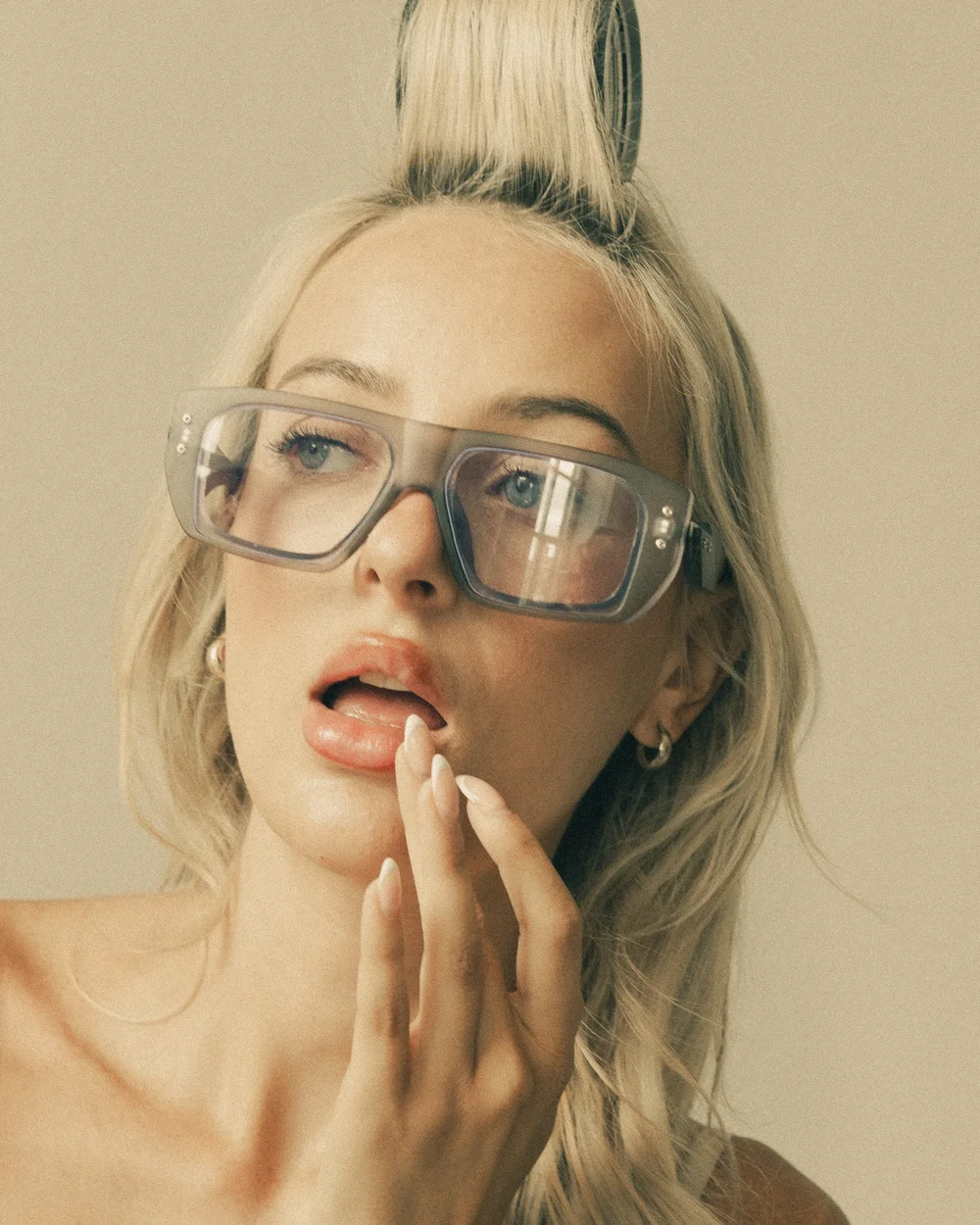 A woman with blonde hair wearing oversized sunglasses, with her tongue out and finger near her lips, against a beige background.