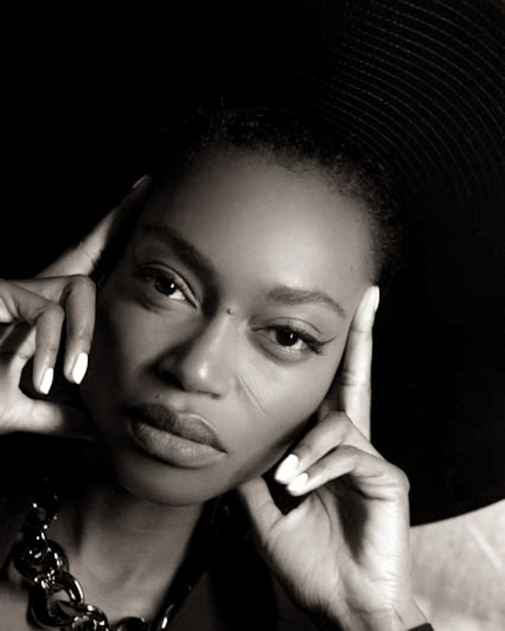 Close-up black and white photo of a woman with dark hair and light skin, smiling, holding her face with her fingers, with part of a black necklace visible.