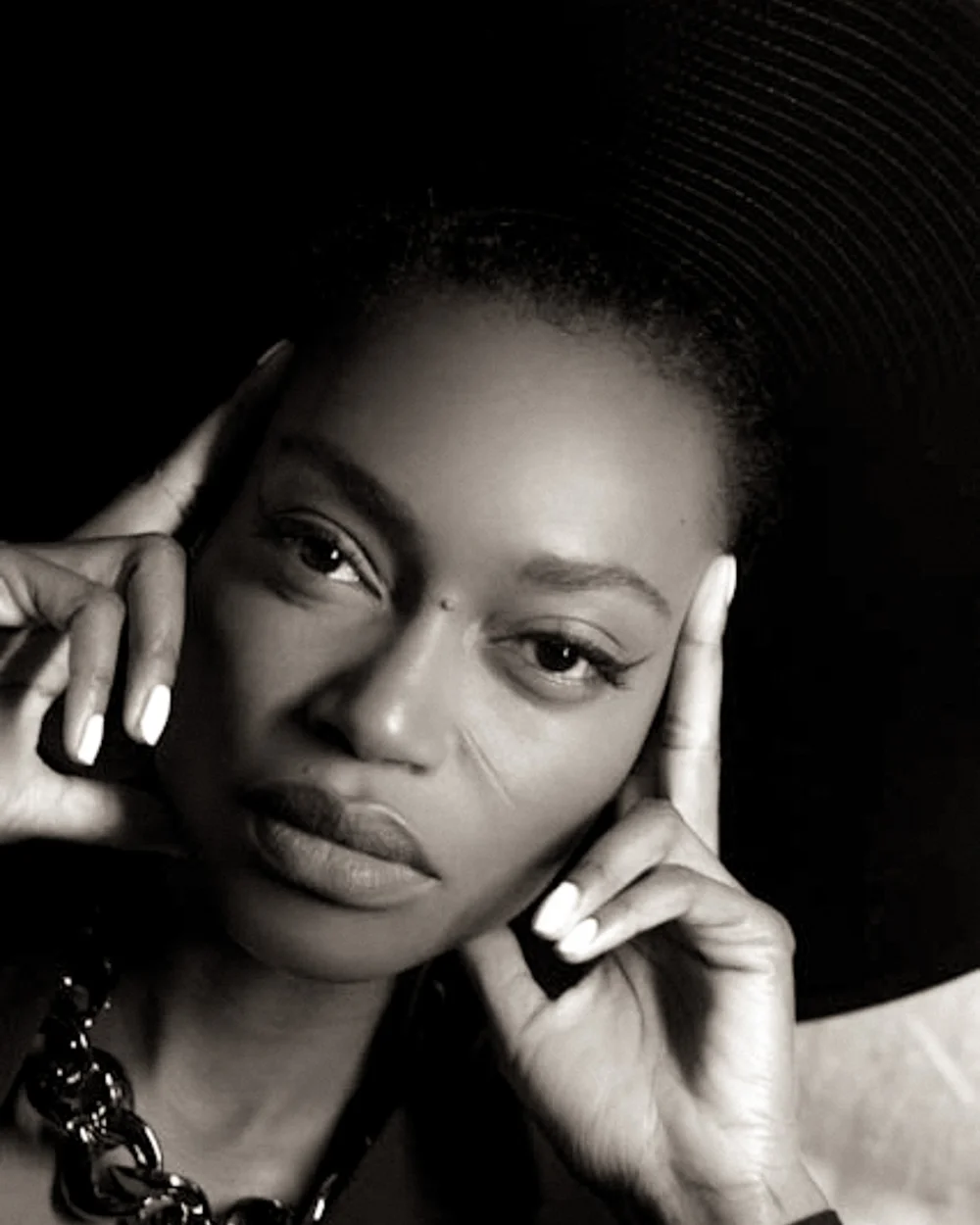 Close-up black and white photo of a woman with dark hair and light skin, smiling, holding her face with her fingers, with part of a black necklace visible.