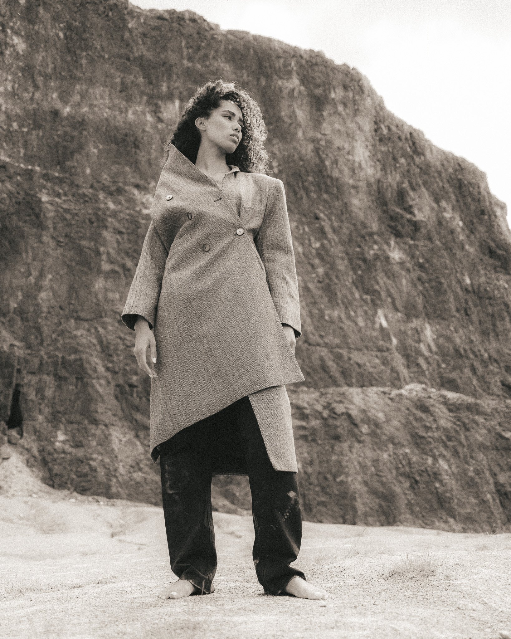 A woman standing on a sandy beach, wearing a coat and black pants, with a rocky cliff in the background.