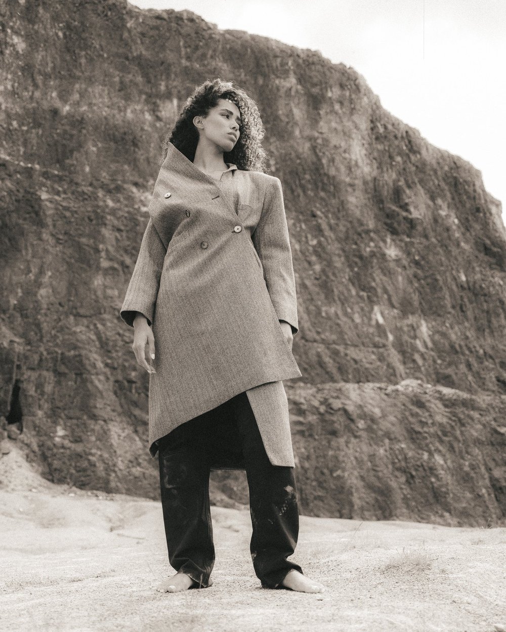 A woman standing on a sandy beach, wearing a coat and black pants, with a rocky cliff in the background.
