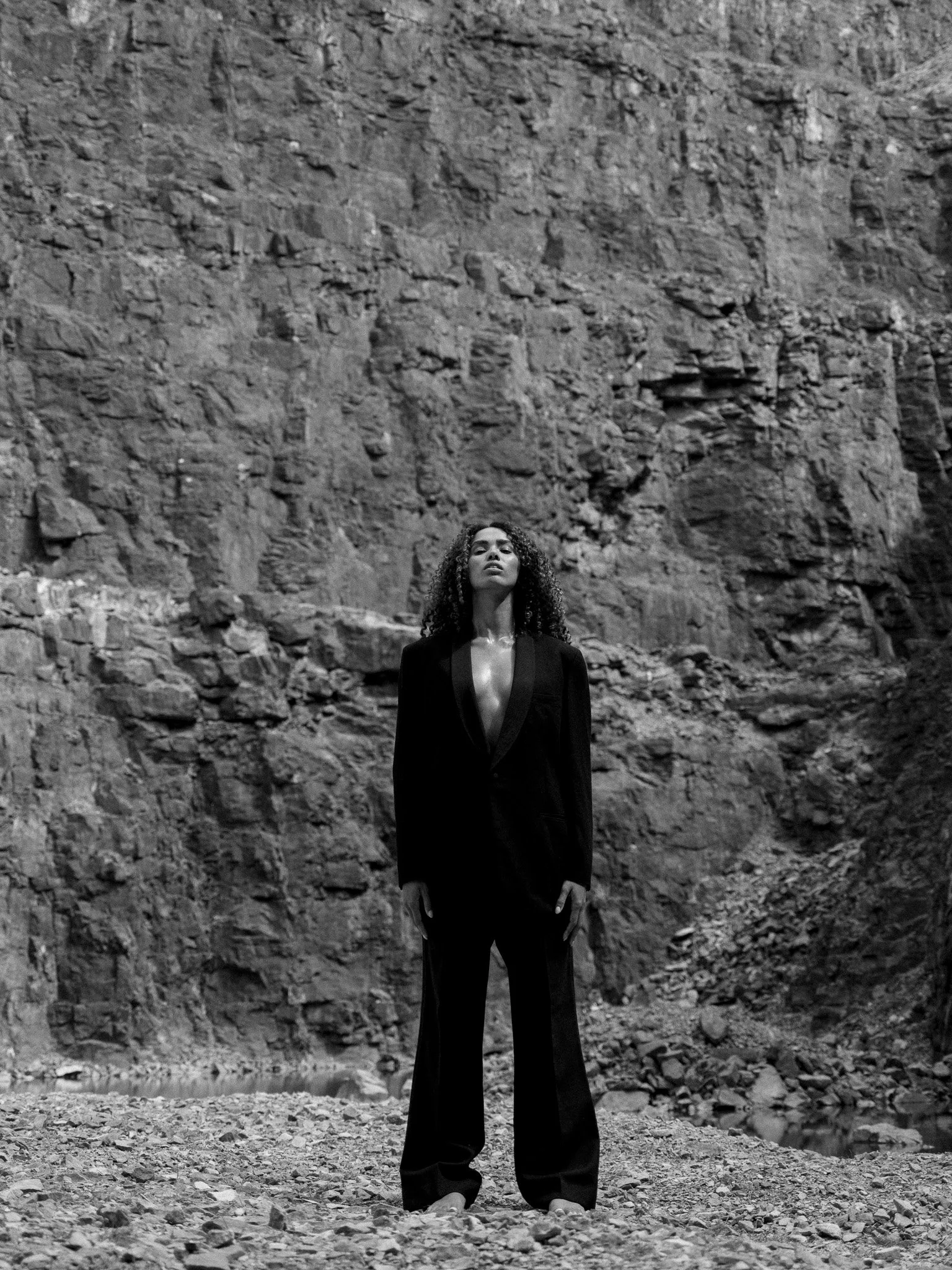 A woman with curly hair standing outdoors in front of a large rocky cliff, wearing a dark blazer and pants, with a serious expression.
