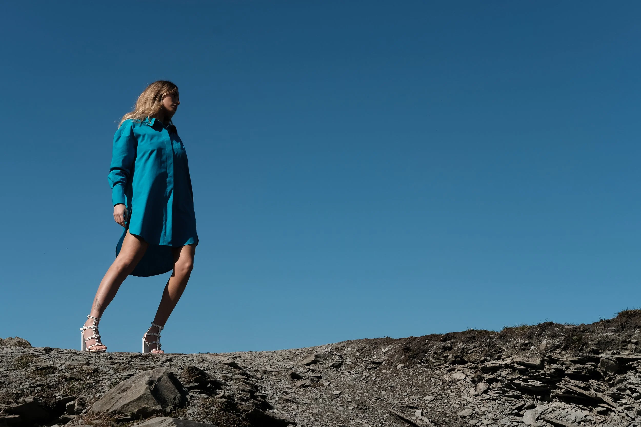 A woman in a bright blue trench coat and white high-heeled sandals standing on rocky terrain under a clear blue sky.
