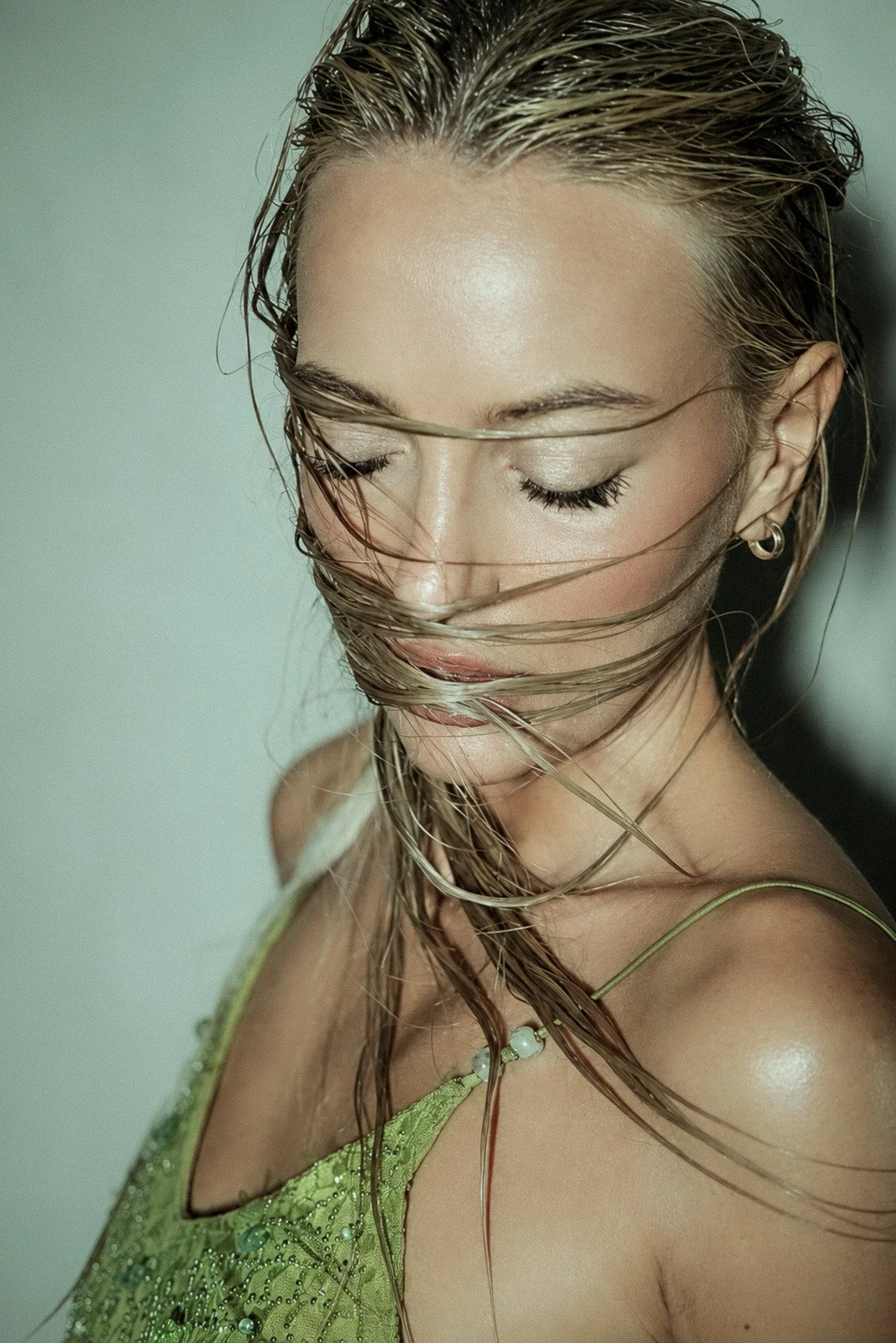 Close-up of a woman with wet hair, wearing a green dress with sparkles and jewelry, with her eyes closed and head slightly tilted.