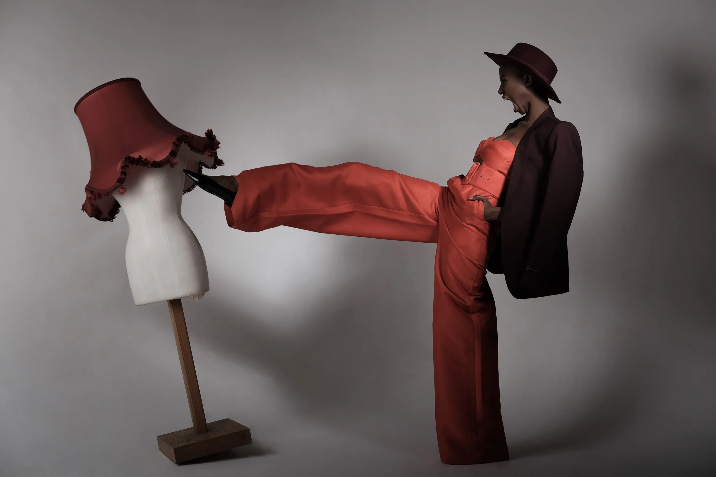 A woman dressed in a bright orange strapless dress with a dark colored jacket and a wide-brimmed hat is kicking an upside-down lampshade that is decorated with a garland of flowers, with her foot.