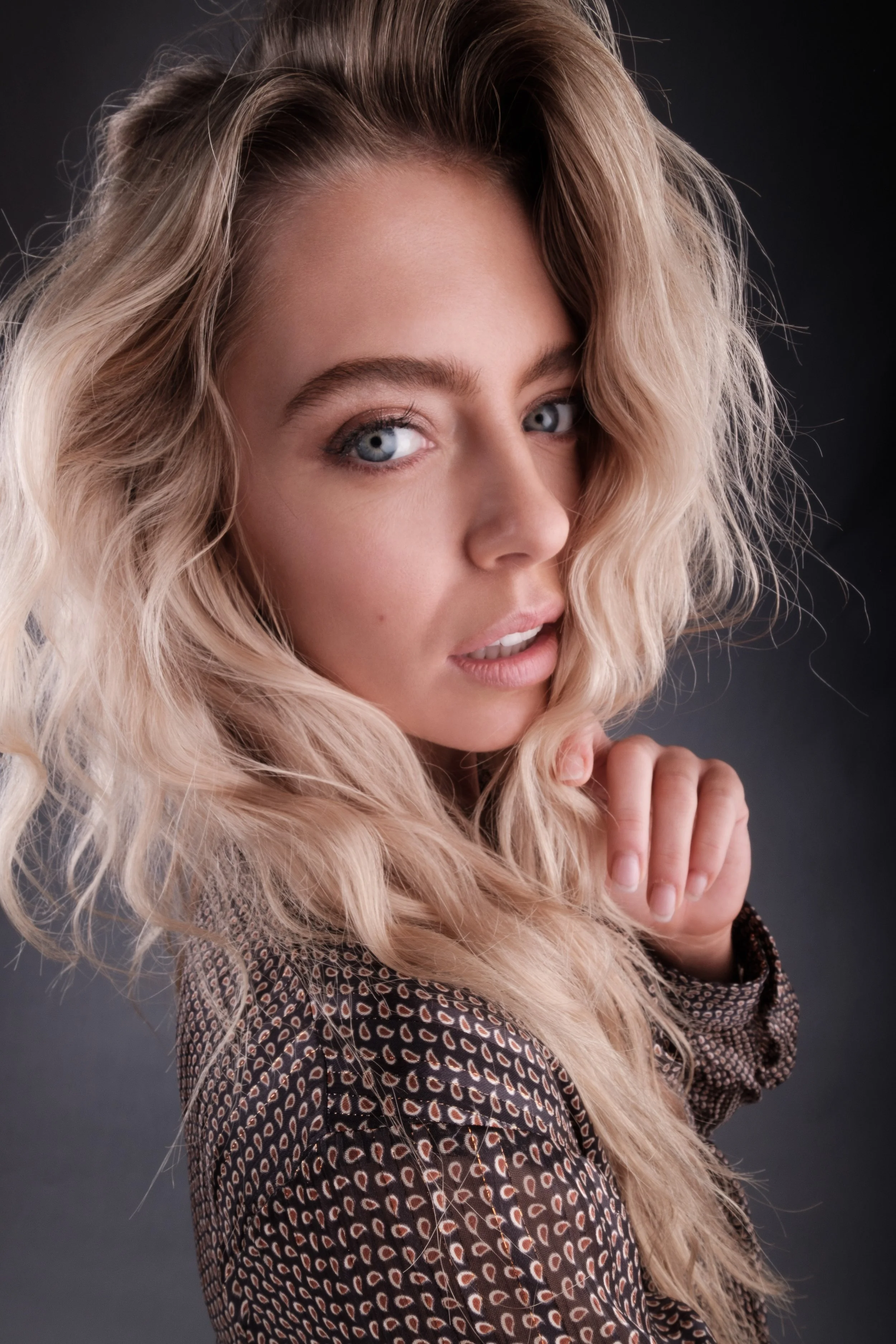 A woman with long, wavy blonde hair and blue eyes, wearing a patterned top, posing with her hand near her face against a dark background.