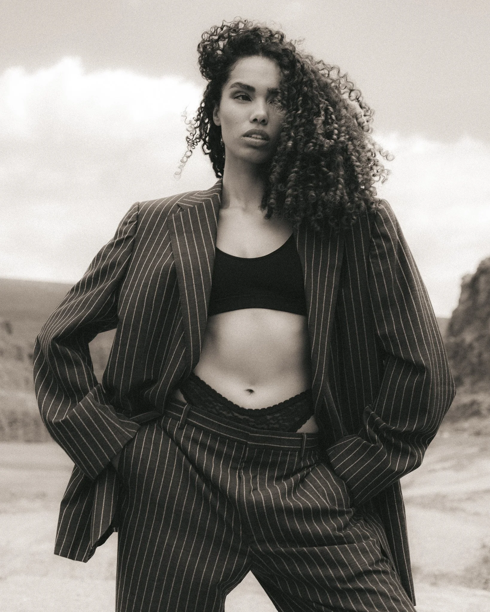 A woman with curly hair wears a pinstripe suit jacket over a cropped top, standing outdoors with her hands in her pockets, in a black-and-white photo.