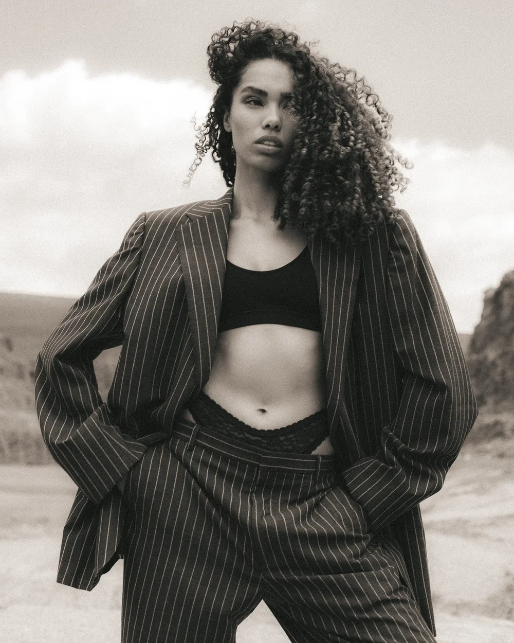 A woman with curly hair wears a pinstripe suit jacket over a cropped top, standing outdoors with her hands in her pockets, in a black-and-white photo.