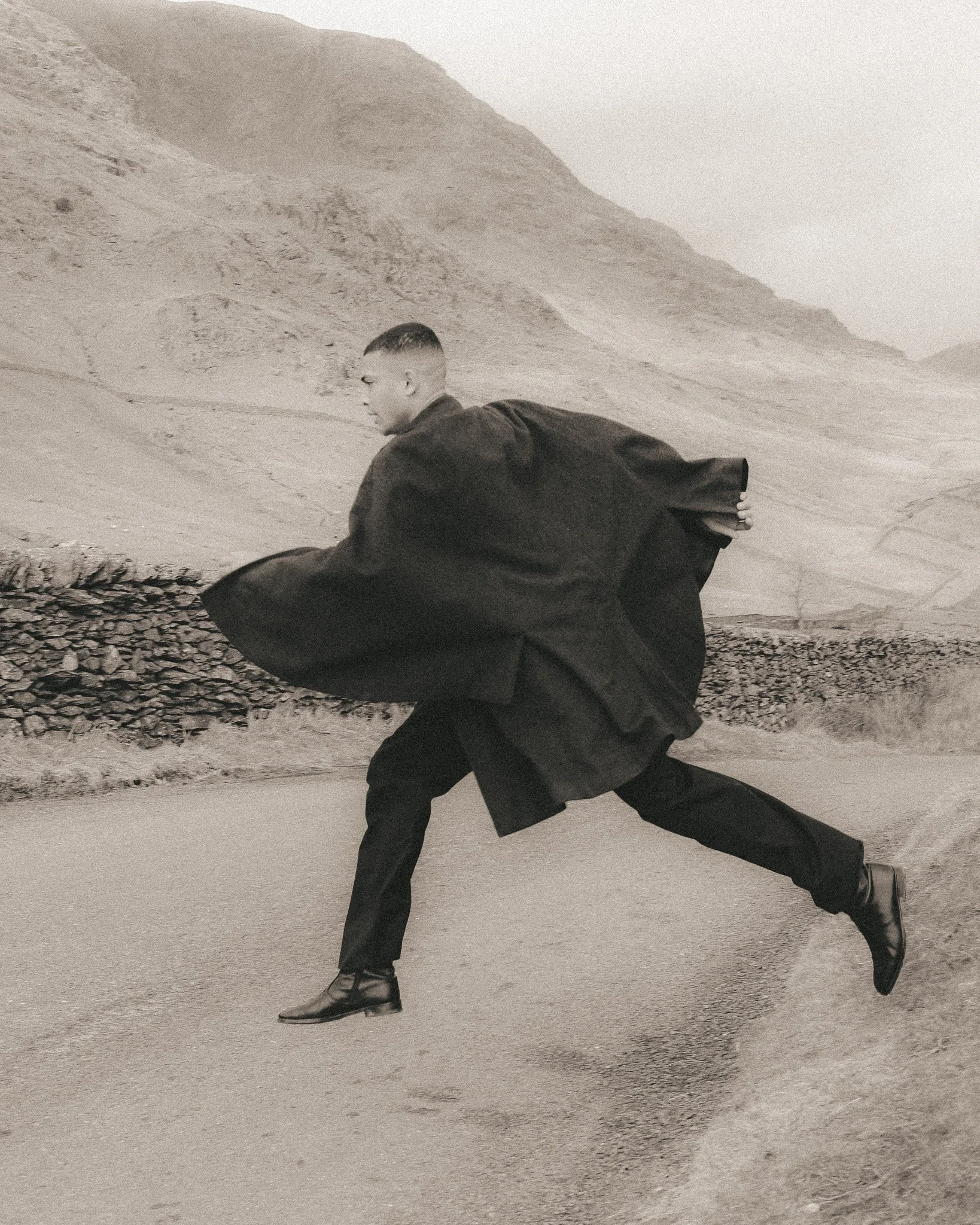 A person wearing a long black coat and black shoes is bent forward with one hand on the ground on a gravel road, in a desert landscape with mountains in the background.
