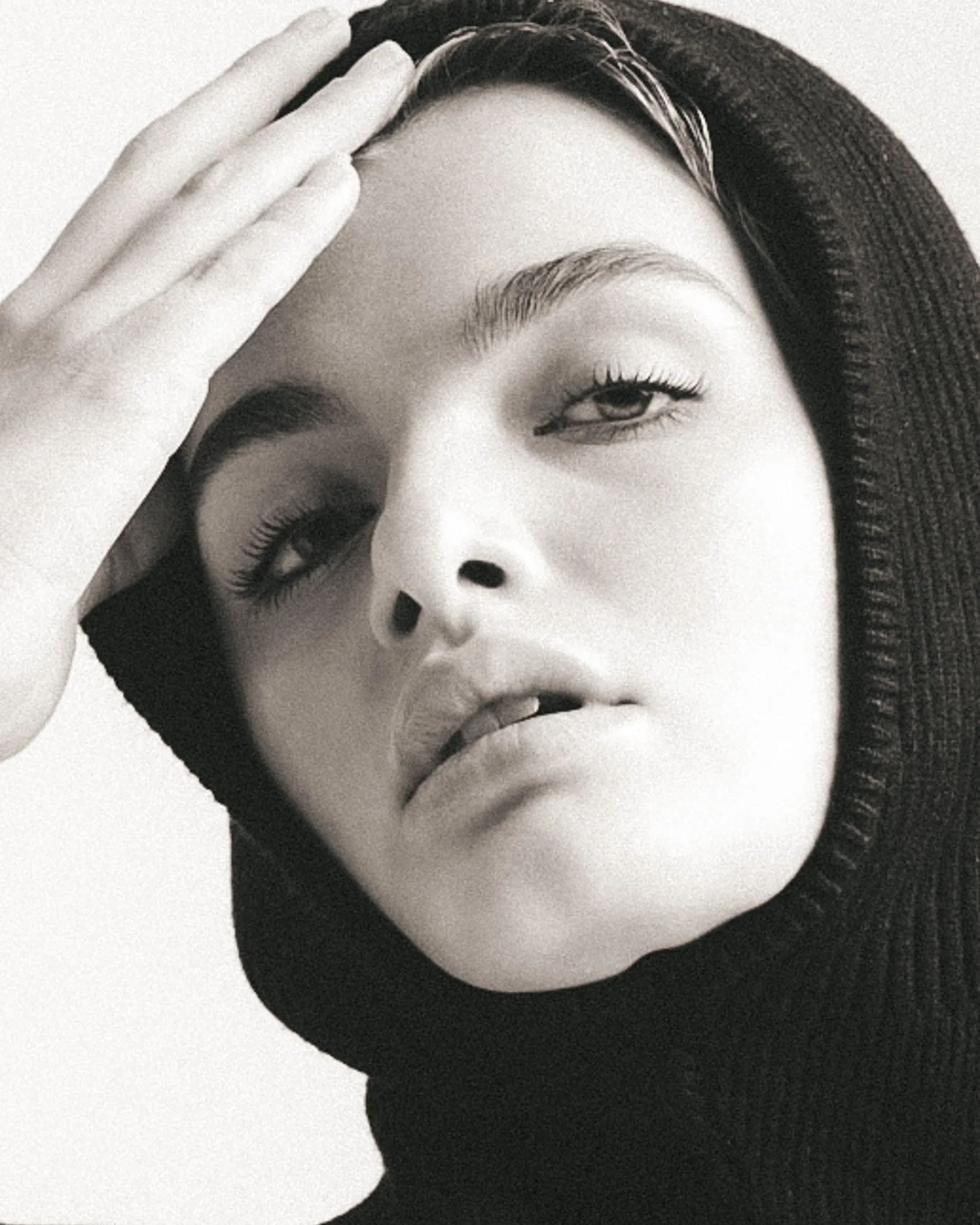A close-up black and white portrait of a woman with smooth skin, well-groomed eyebrows, long eyelashes, subtle eye makeup, and full lips. She is wearing a dark hat, and her hair is tucked underneath it. Her hand is resting on her forehead.