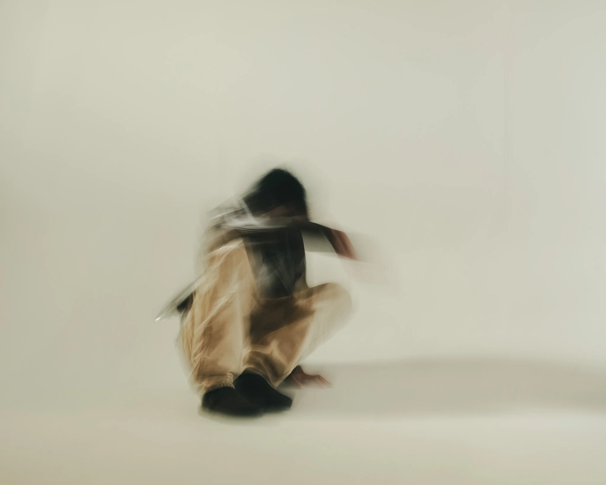 Blurred image of a person walking on a plain white or light-colored wall background, wearing a jacket, pants, and black shoes.
