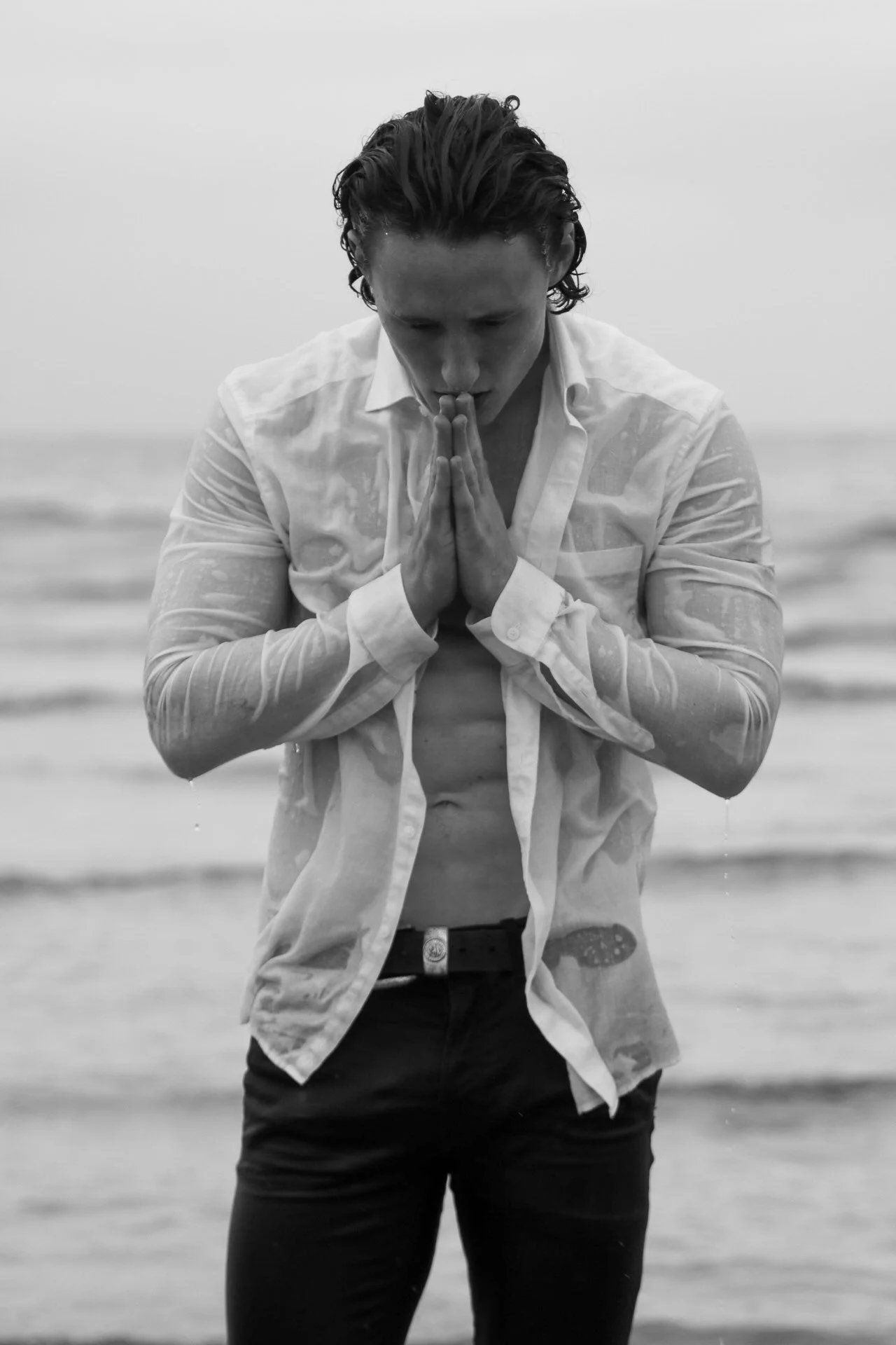 Black and white photo of a man standing on the beach, praying with hands pressed together in front of his face, wearing a partially unbuttoned white shirt and black pants.