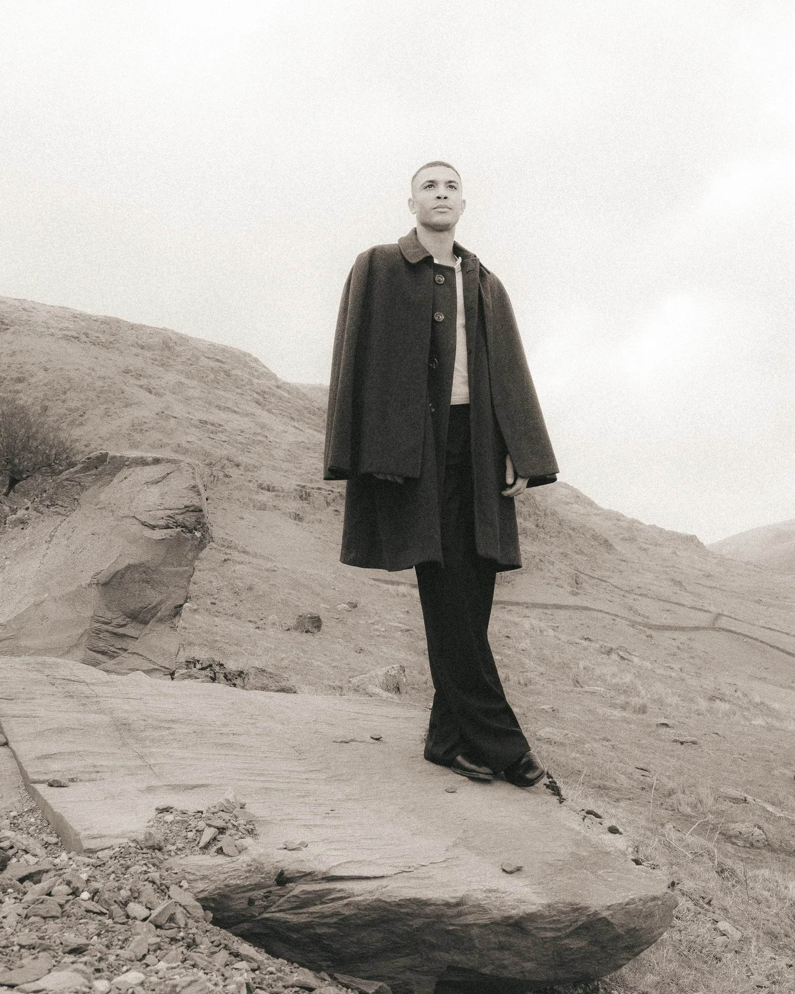 A person with short hair wearing a dark coat and long pants standing outdoors on rocky terrain with hills in the background, looking upward.
