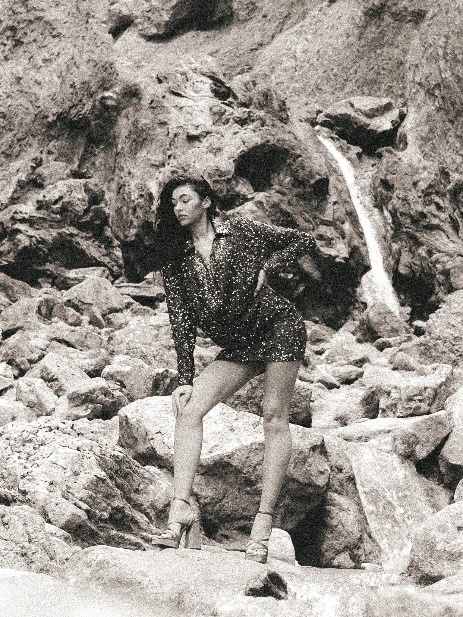 A woman with long dark curly hair poses among rocks and boulders, wearing a sequined dress and high heels.