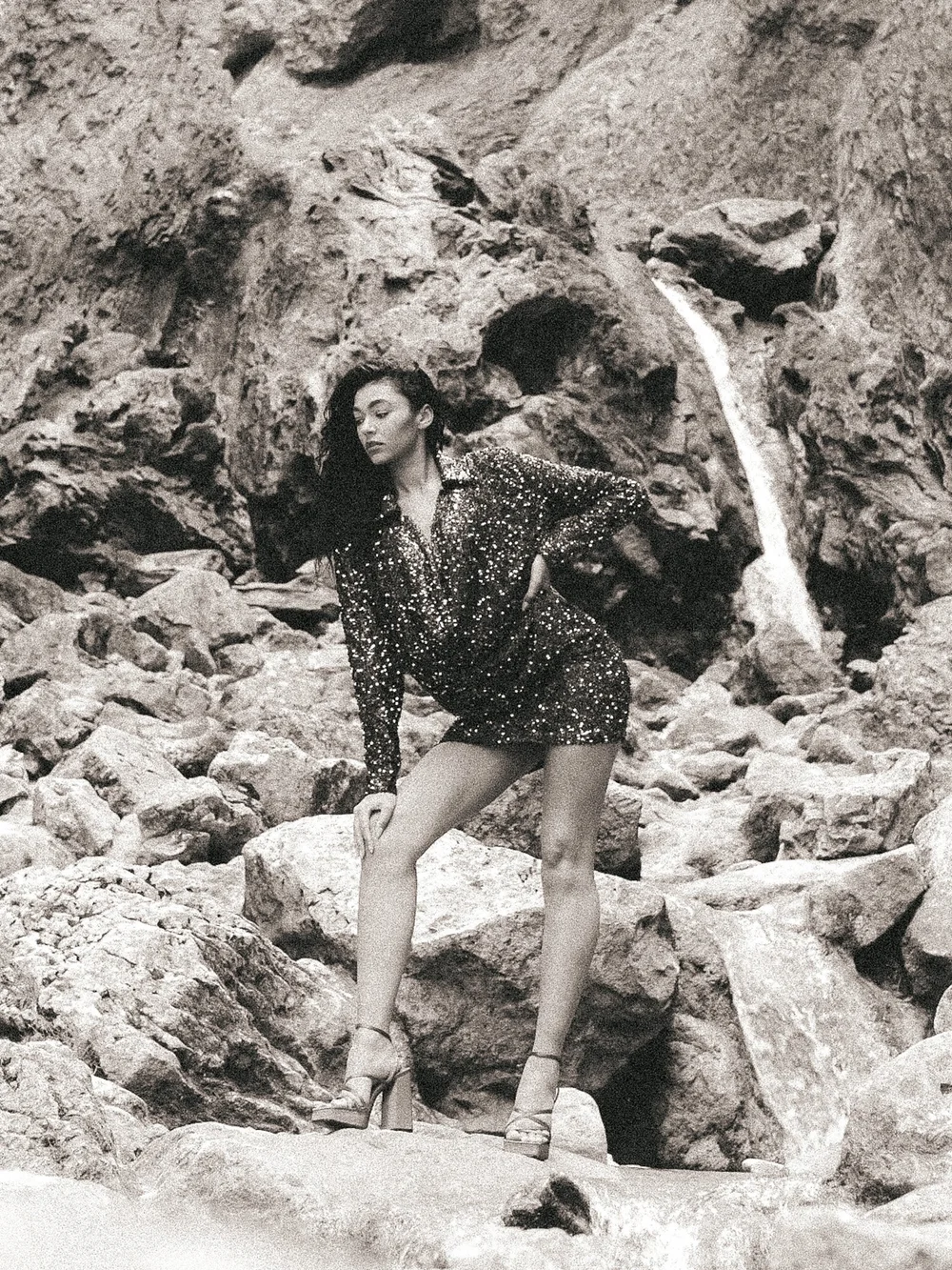 A woman with long dark curly hair poses among rocks and boulders, wearing a sequined dress and high heels.