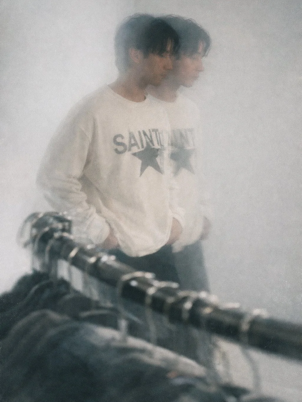 A person with dark hair standing in a foggy environment, wearing a white Saint Laurent sweatshirt, with a motorcycle or bicycle in the foreground.