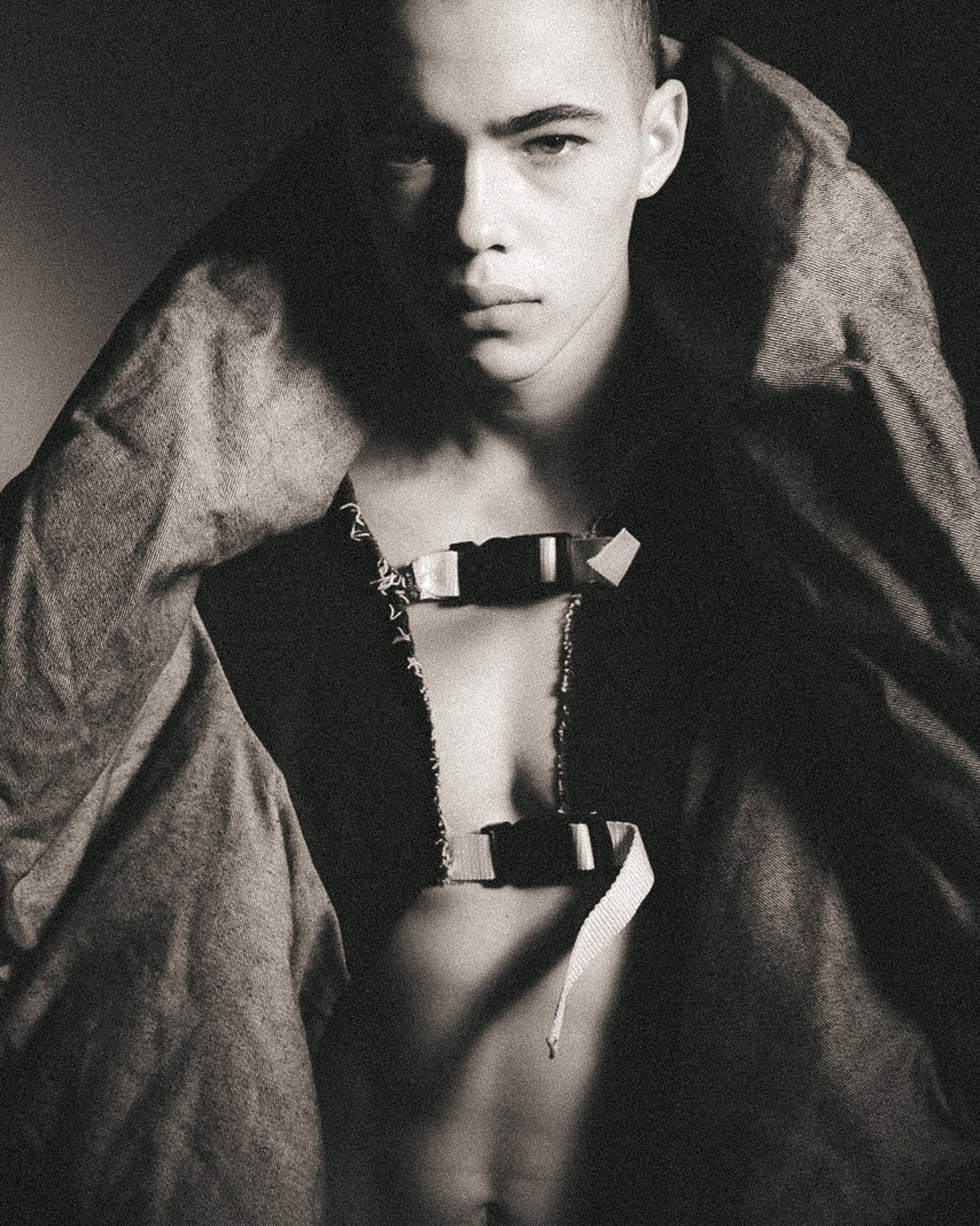 Black and white close-up of a person with short hair, wearing a jacket and a necklace, with a serious expression, looking directly at the camera.