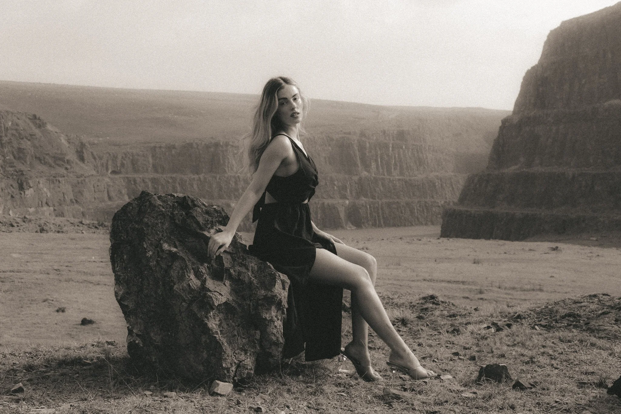 A woman sitting on a large rock in a desert landscape with cliffs in the background, wearing a black dress.