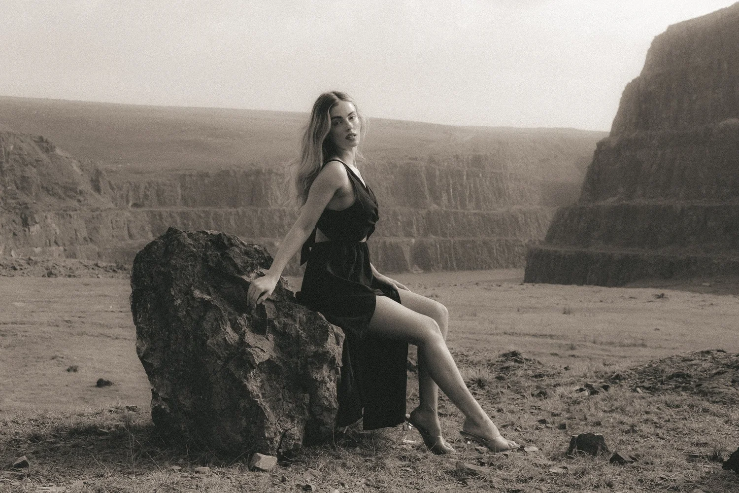 A woman sitting on a large rock in a desert landscape with cliffs in the background, wearing a black dress.