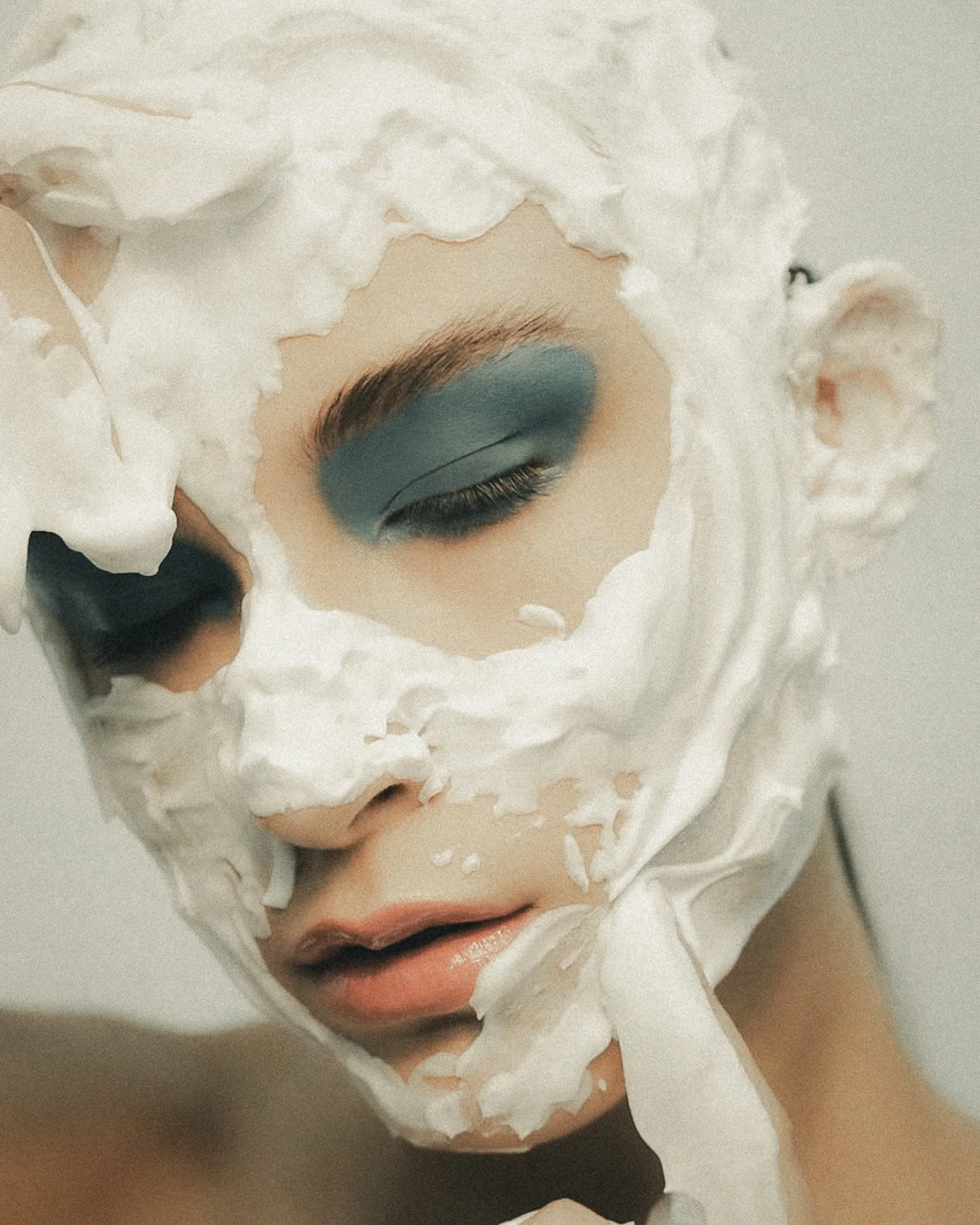 Close-up of a person's face with closed eyes, wearing eye makeup, partially covered with torn white tissue paper.