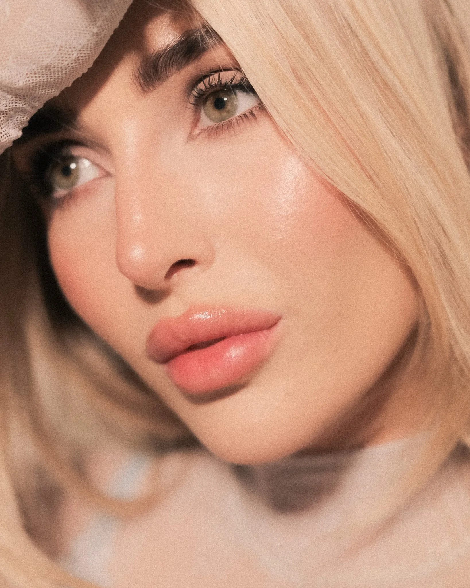 A close-up of a woman's face, showing her brown eyes, full lips, and a portion of her blonde hair, with part of her face hidden behind her hand.