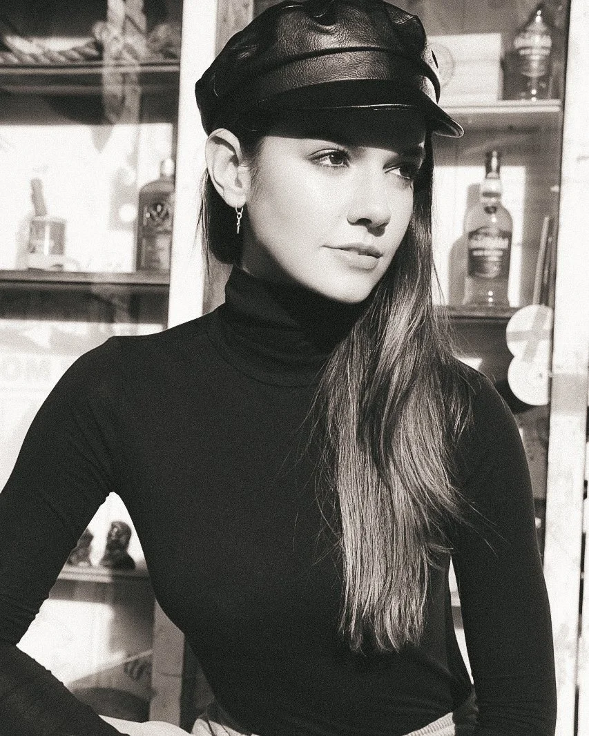 A young woman with long hair wearing a black turtleneck, a leather cap, and earrings. She is looking to the side, indoors with shelves and bottles in the background.