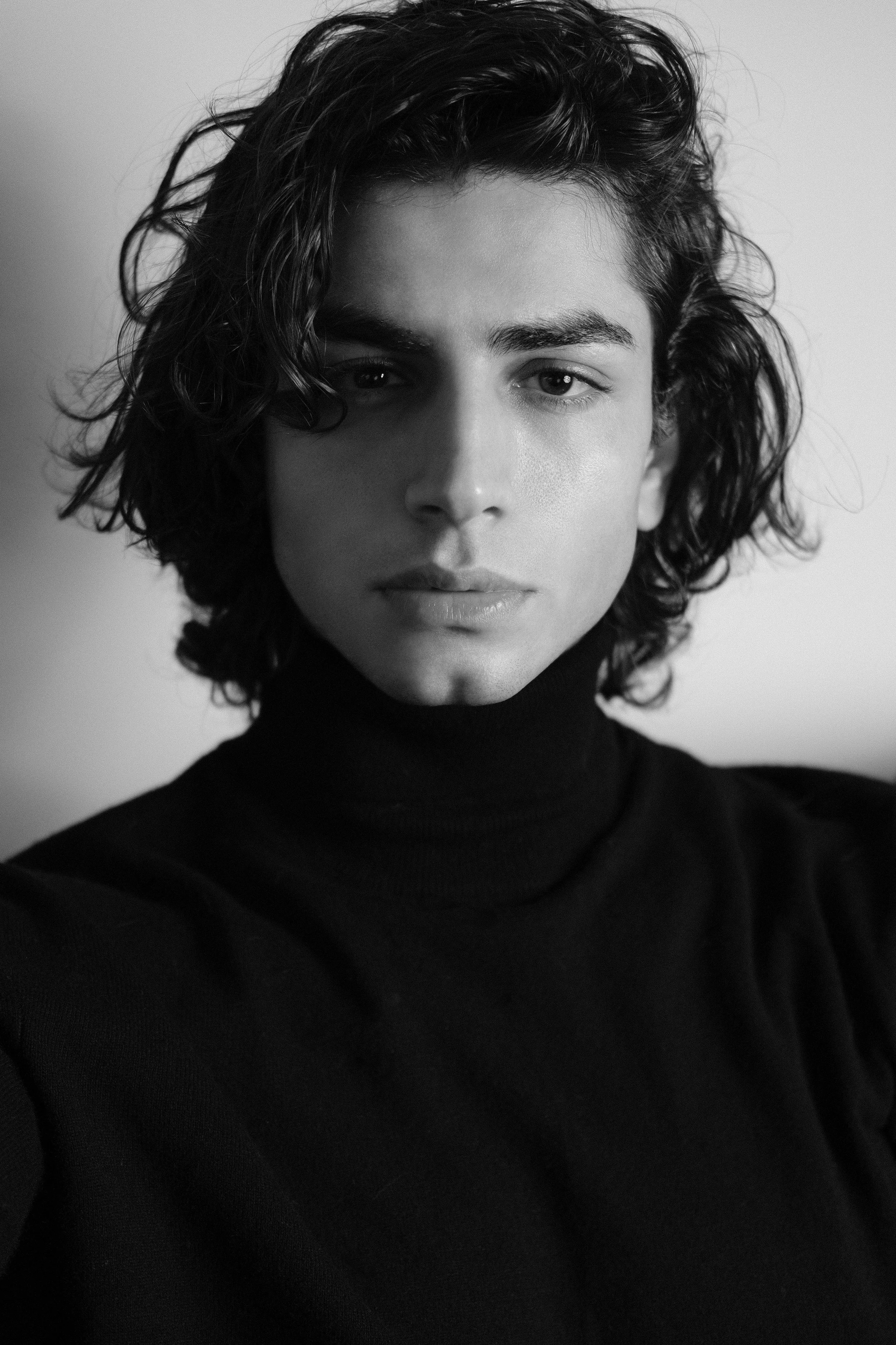 A black and white close-up portrait of a young person with wavy, shoulder-length hair, wearing a turtleneck sweater, looking directly at the camera with a neutral expression.