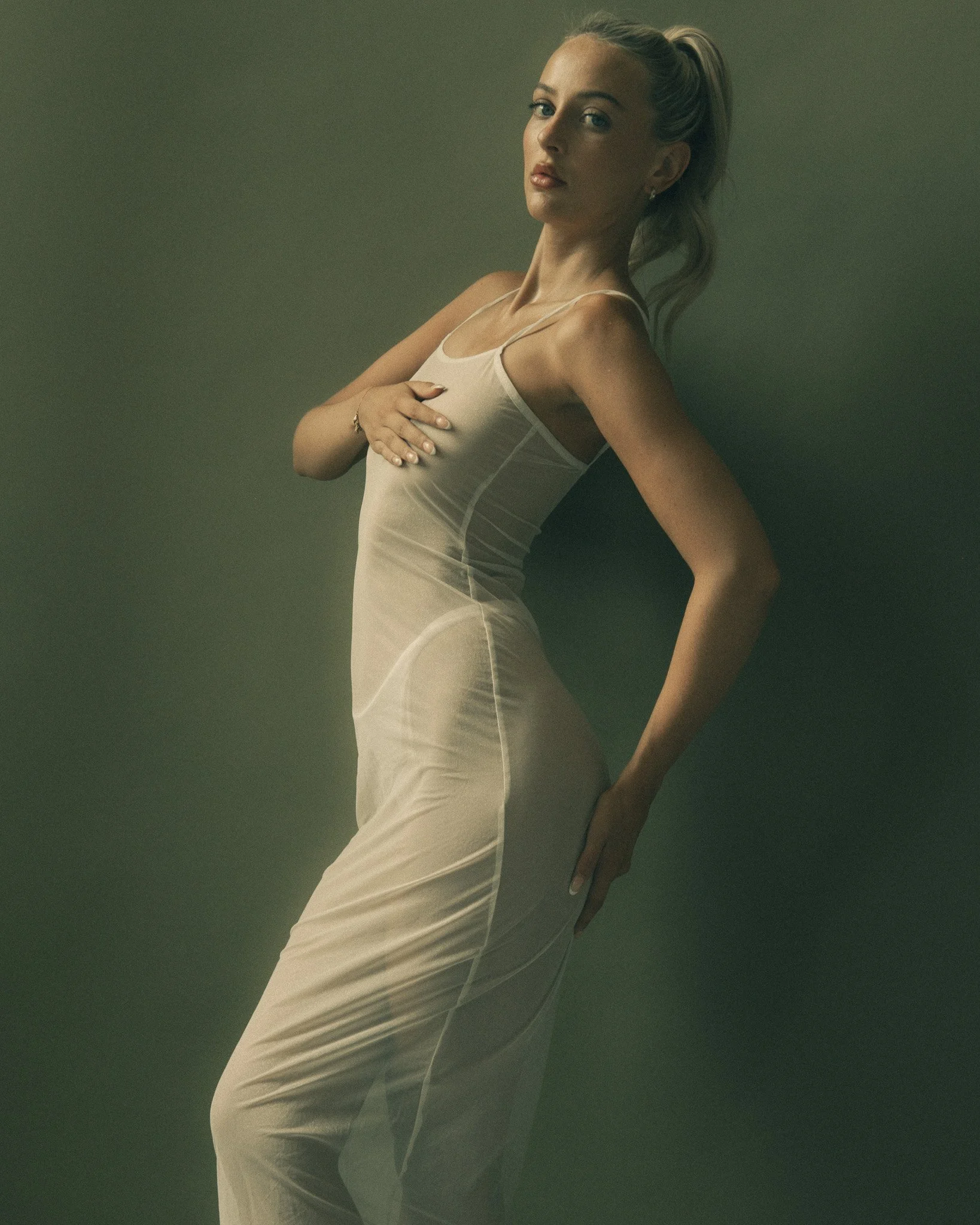A woman with blonde hair tied in a ponytail is wearing a fitted, sleeveless beige dress and jewelry, posing in front of a plain, dark green background.