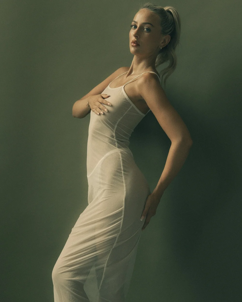 A woman with blonde hair tied in a ponytail, wearing a white sleeveless dress, standing against a greenish background with a neutral expression.