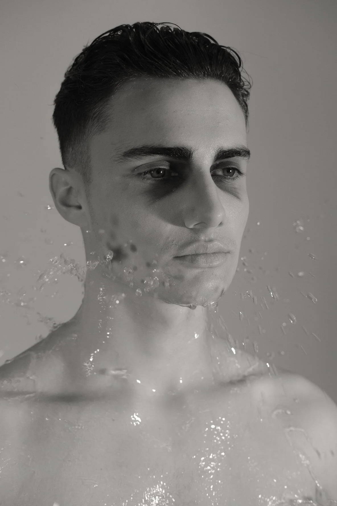 Black and white photo of a young man with styled hair, minimal makeup, and a serious expression, submerged in water up to his shoulders, with water splashing around him.