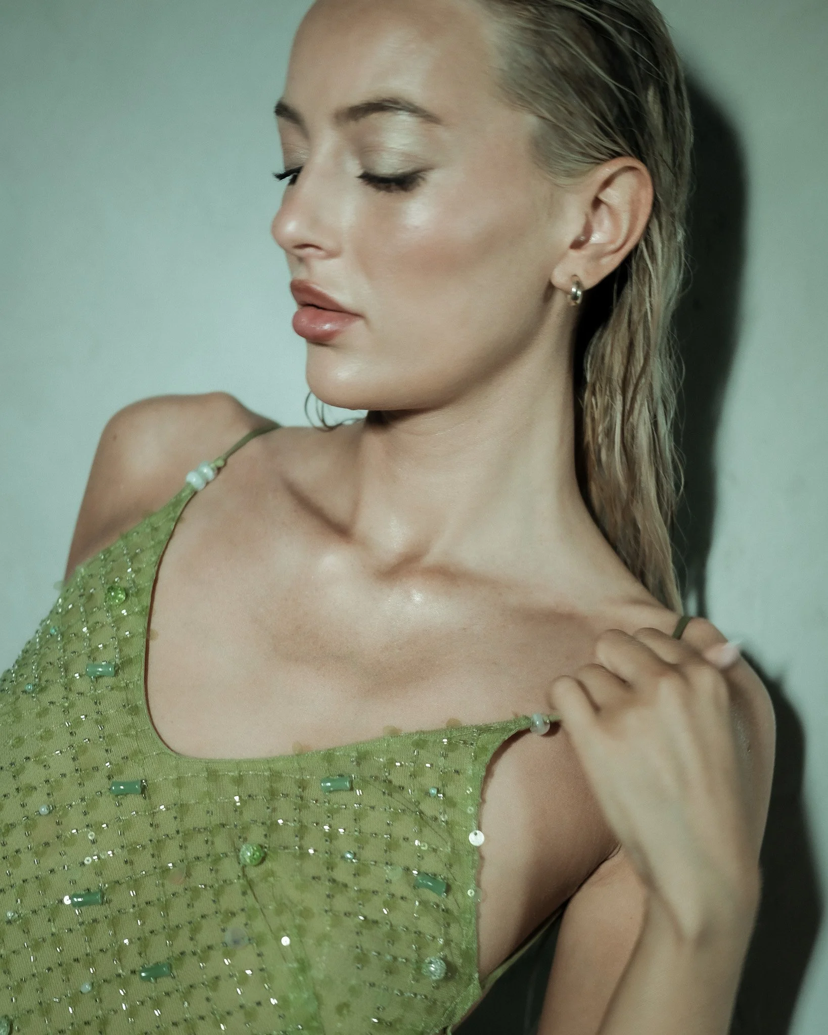Close-up of a woman with fair skin and blonde, wet hair, wearing a light green, beaded dress with spaghetti straps and small hoop earrings, standing with her eyes closed in front of a plain wall.